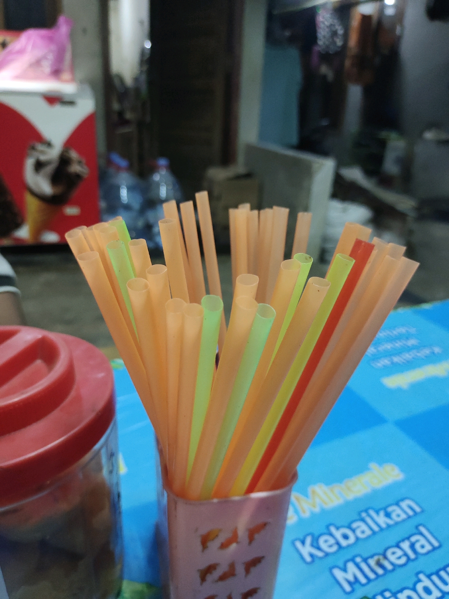 A close-up, slightly blurry, eye-level outdoor shot shows a bunch of colorful plastic drinking straws in a pink cup on a blue table. The straws are predominantly orange, with some green and one red straw interspersed. To the left, partially visible, is a clear jar with a red lid, containing food items. In the background, out of focus, are various objects including a red and white refrigerator, plastic bottles, and what appears to be an outdoor seating area with more tables and chairs. The lighting suggests it is daytime. This scene is likely from Lengkong Kulon, Indonesia, given the context provided.