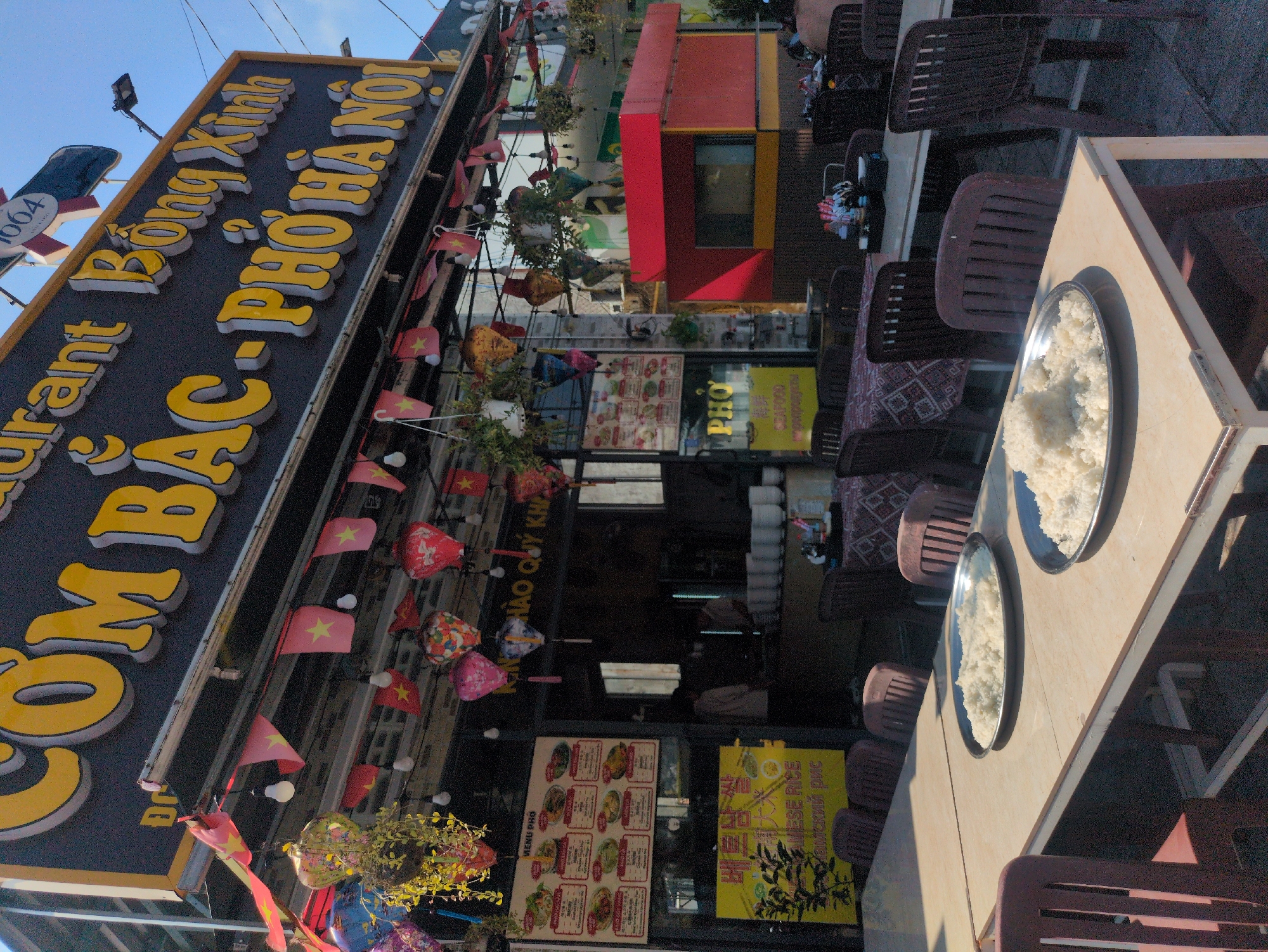 The image captures an outdoor scene at a restaurant called "Cơm Bắc - Phở Hà Nội" (Northern Rice - Hanoi Pho) in Phú Quốc, Vietnam. The restaurant's storefront is decorated with Vietnamese flags and lanterns. Menu boards with images of food items are displayed on the glass doors. The interior is partially visible, with stacks of bowls and possibly servers. In the foreground, a table is set with plates of rice. Brown plastic chairs surround the table.