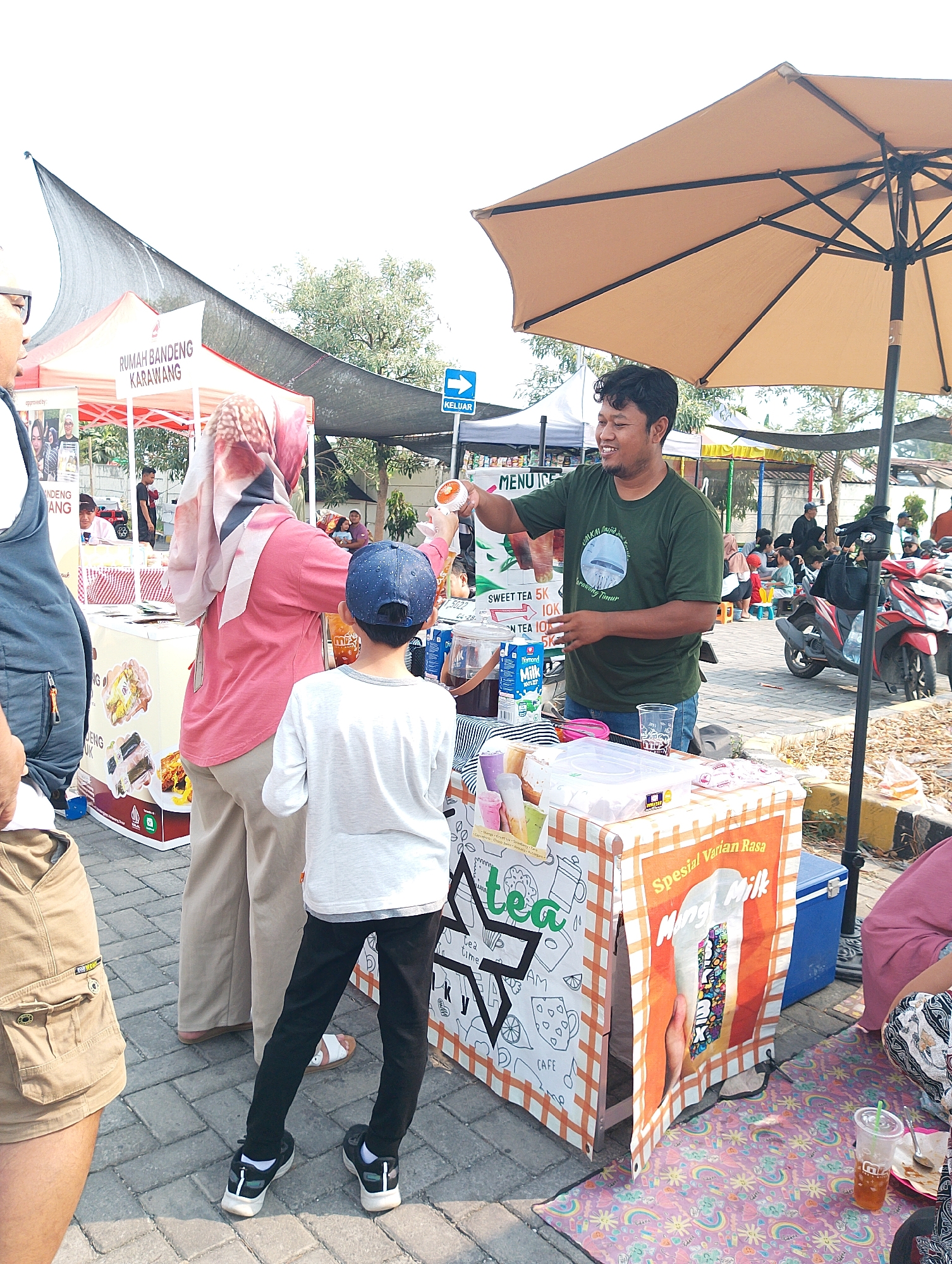 This image depicts an outdoor market scene in Karawang, Indonesia, likely a food stall event. In the foreground, a vendor wearing a green t-shirt smiles and appears to be serving a drink from a stand. The stand has colorful signage, with one panel displaying text including "Spesial Varian Rasa" and a picture of a drink. Next to the stand, there is an open cooler.
Several people are visible around the stall. A woman in a pink shirt and headscarf stands to the left of the vendor, holding a small fan. A young boy in a blue baseball cap and white shirt stands facing the vendor. Other individuals are in the background, including some seated and others standing near parked motorcycles.
In the middle ground, a red tent with "RUMAH BANDENG KARAWANG" written on it is visible, suggesting a connection to a local establishment. The ground is paved with cobblestones, and an umbrella provides shade over the vendor's area. The overall environment suggests a sunny day at a lively outdoor gathering.