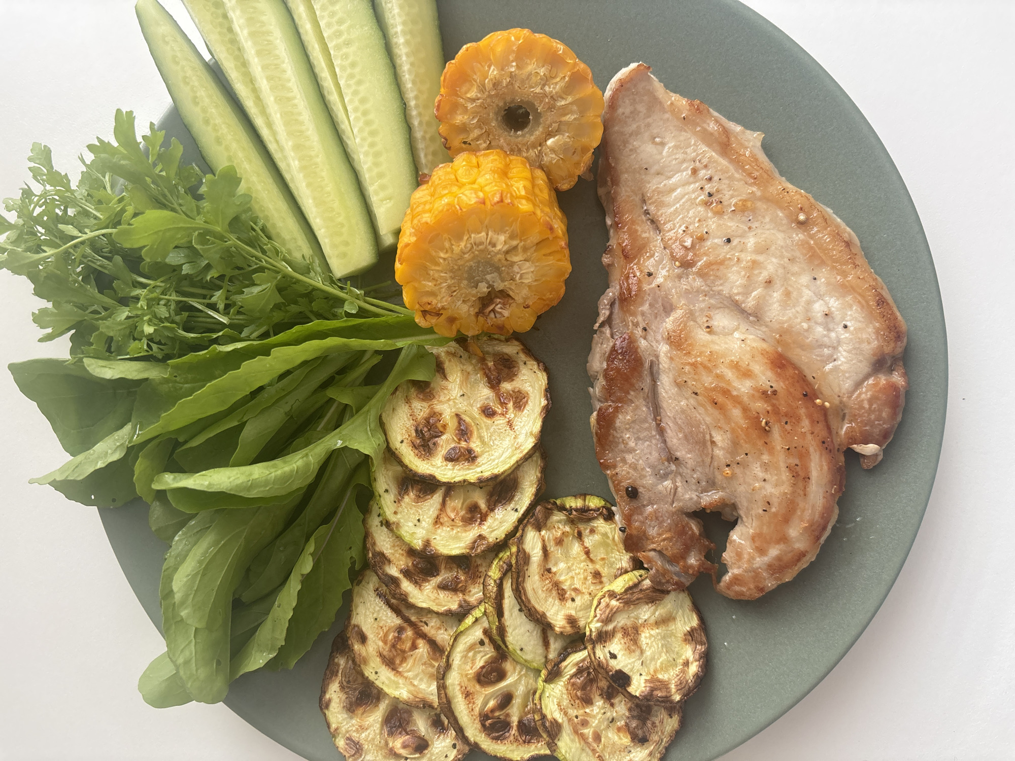 A close-up, overhead shot displays a grey plate filled with food. On the bottom left of the plate, a large piece of cooked meat, possibly chicken or turkey breast, is seasoned with salt and pepper. To its left are several slices of grilled zucchini, charred in places. Above the zucchini and to the right of the meat are two circular slices of corn on the cob. A bundle of fresh green leafy vegetables, likely arugula and parsley, sits at the top of the plate, with several sliced cucumbers to their right. The background is a plain white surface. There are no people or animals visible in the image. The scene is indoors and the lighting is bright and natural. The location in Chaikovsky, Russia, cannot be determined from the visual information provided in the image.