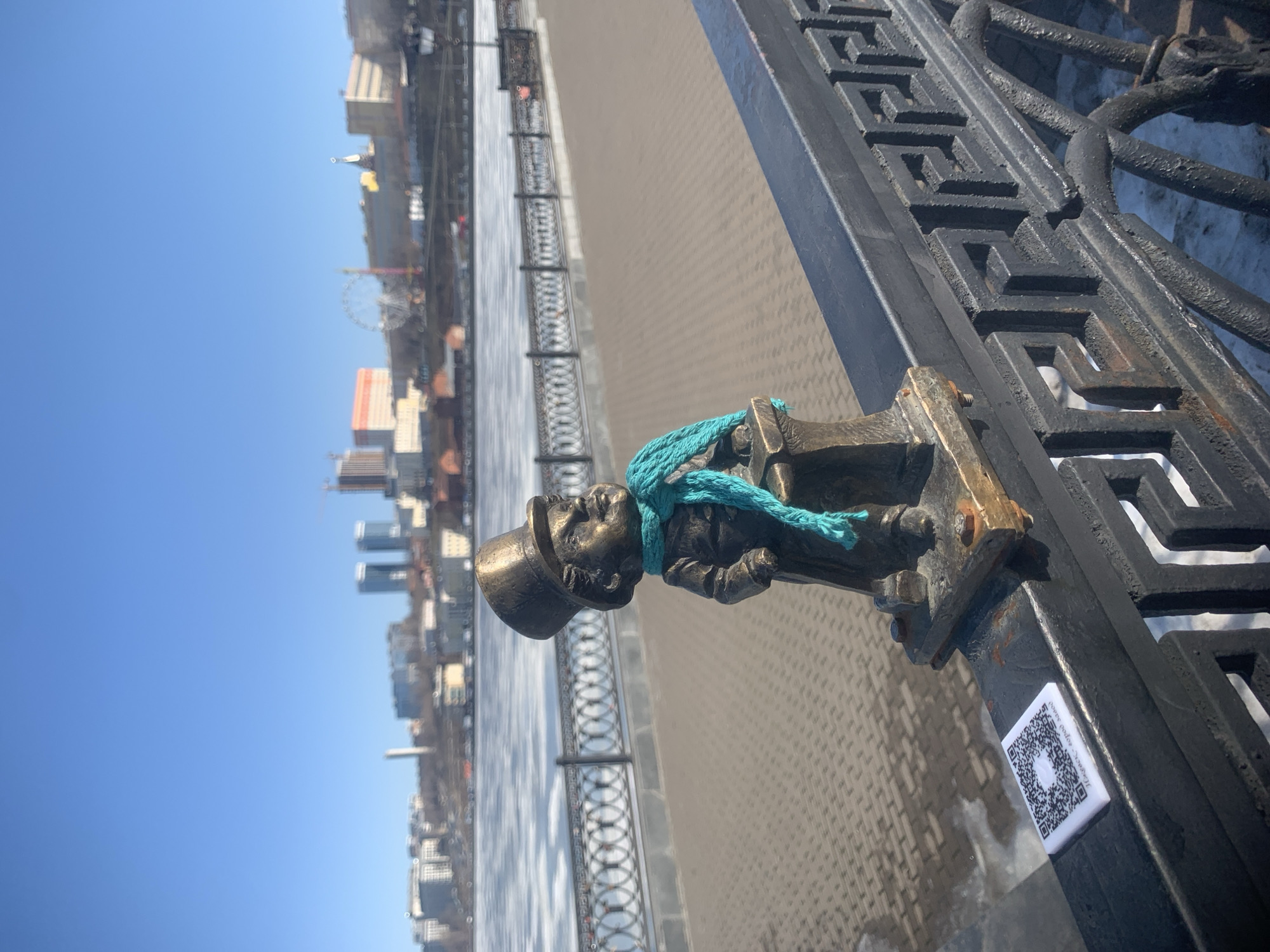 The photograph presents a close-up, slightly angled view of a bronze statue of a small man in a top hat and a turquoise scarf. The statue is affixed to a black metal railing. In the background, out of focus, is a cityscape including buildings, a ferris wheel, and a waterfront. A QR code sticker is visible near the statue's base on the railing. The location is Izhevsk, Russia.