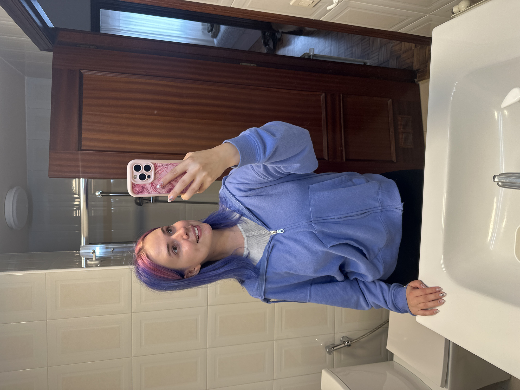 A young woman with dyed purple and pink hair takes a selfie in a bathroom. She is wearing a light blue zip-up hoodie over a white striped shirt. She holds a pink cell phone in her right hand. Her left hand rests on a white sink. The bathroom features cream-colored, square-tiled walls. Behind the woman is a dark wood cabinet. The location is a bathroom in Gijón/Xixón, Spain.
