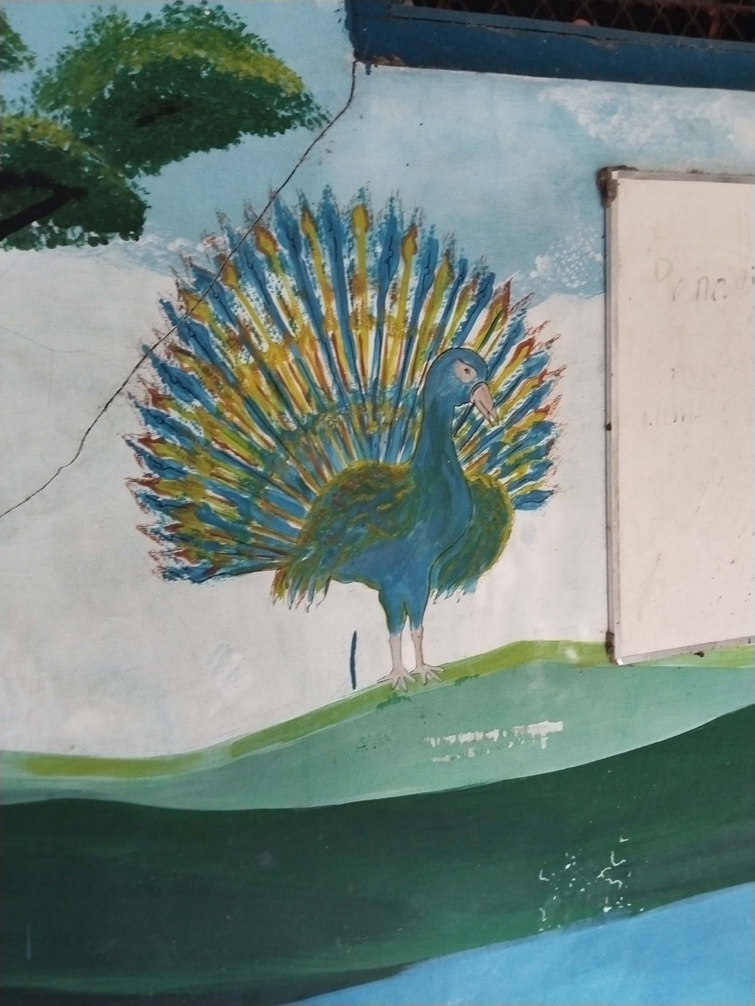 A painting of a peacock on a wall. The peacock is blue with a yellow and blue fan of feathers. The background is a light blue sky with a green tree on the left. The peacock is standing on a green hill. There is a white board on the right side of the painting. This image was taken in Guworejo, Indonesia.
