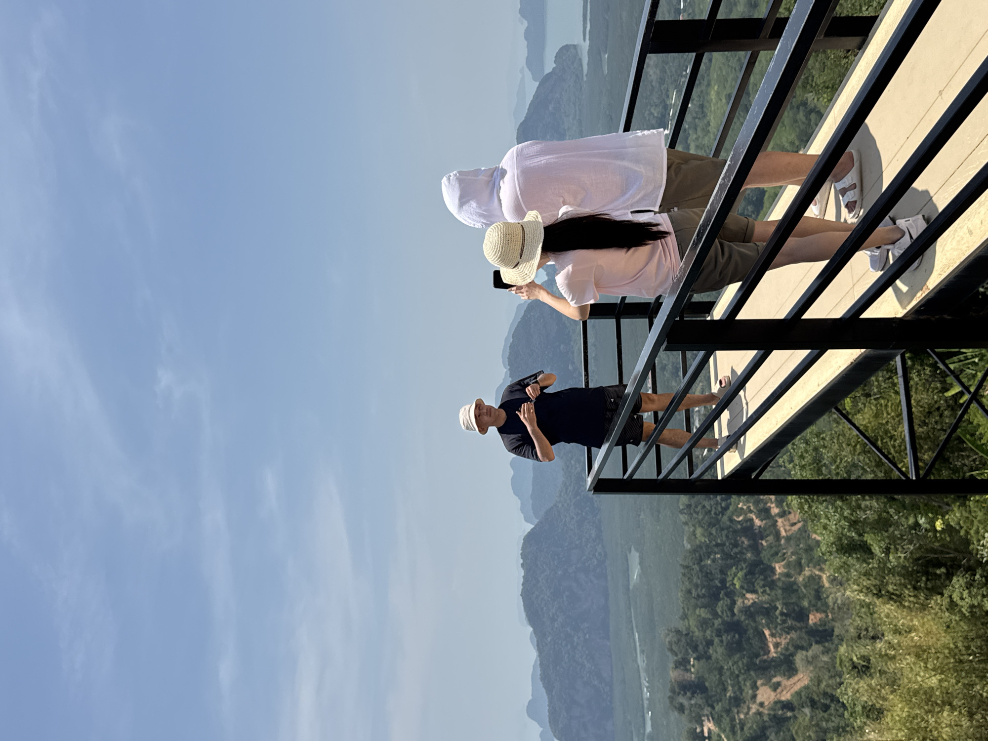 The image shows a group of three people standing on a lookout platform in Ban Khlong Khian, Thailand. The platform has a black metal railing and a light-colored wooden floor. The view from the platform includes mountains, trees, and water in the distance. One person is wearing a black shirt, black shorts, and a light-colored hat. Another person is wearing a white hooded shirt. The third person is wearing a white hat and is holding a phone, presumably taking a picture. They are all observing the landscape.