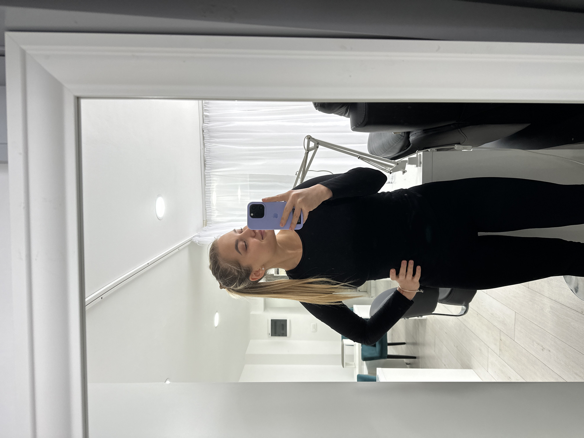 A woman stands in front of a large, rectangular, white-framed mirror taking a selfie with a lavender-colored smartphone. She is wearing a black, long-sleeved top and black leggings. Her blond hair is pulled back into a low ponytail. The room has white walls, a white ceiling with recessed lighting, and light-colored wood flooring. In the background are several chairs, including a black reclining treatment chair, and what appears to be a white treatment table with an adjustable lamp. The location may be a salon or clinic in Budva, Montenegro, but this cannot be determined definitively from the image.