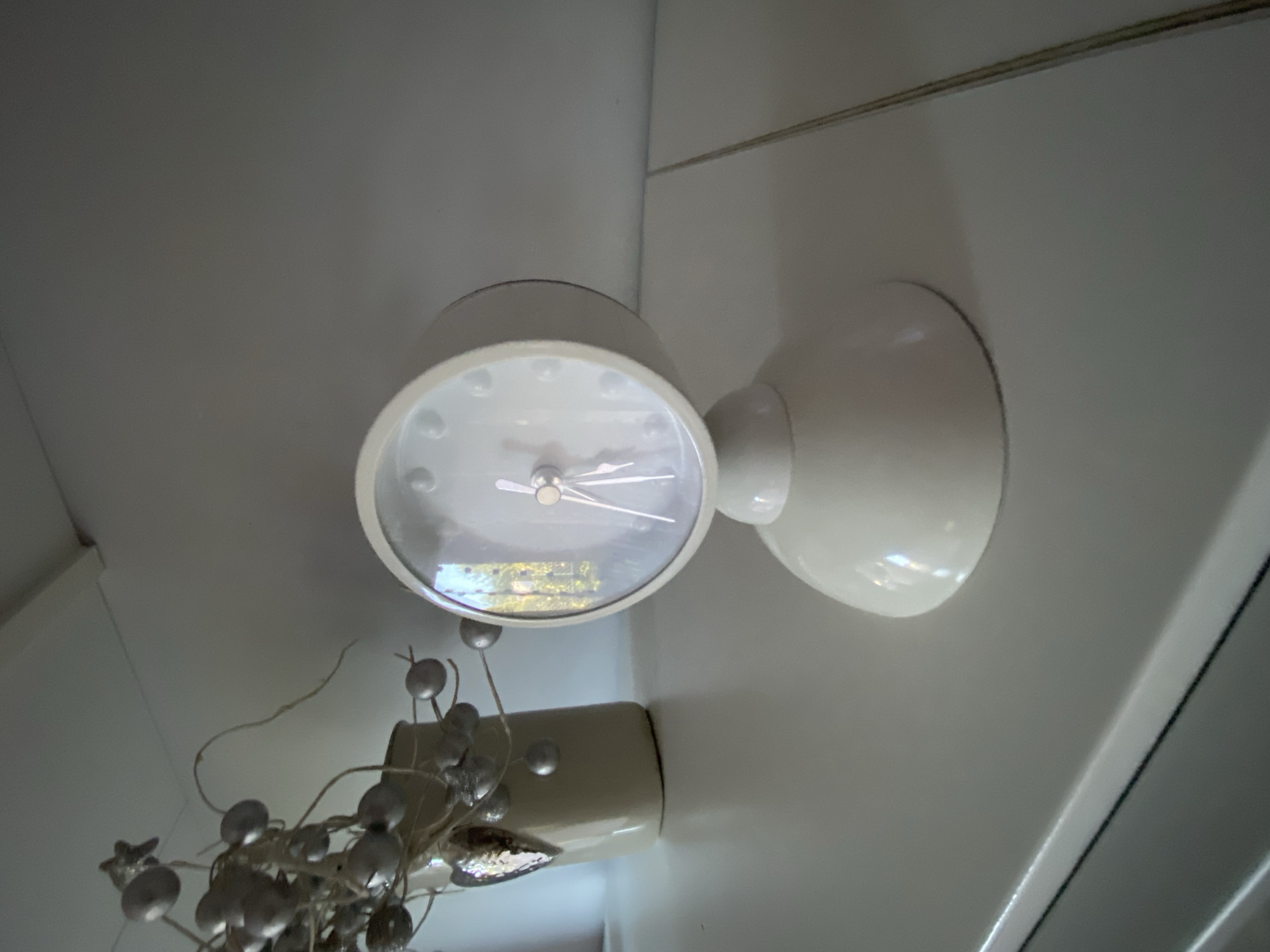 A light-colored, retro-style alarm clock with a round face and a domed base sits on a white surface. The clock face has no numbers, only raised dots for the hours and slender silver hands. To the left of the clock, a cream-colored vase holds decorative branches adorned with silver baubles and a metallic heart-shaped ornament. The objects are placed on a white shelf or table against a plain, light-colored wall. This scene does not provide any information that would allow for a specific location in Wrocław, Poland to be determined.