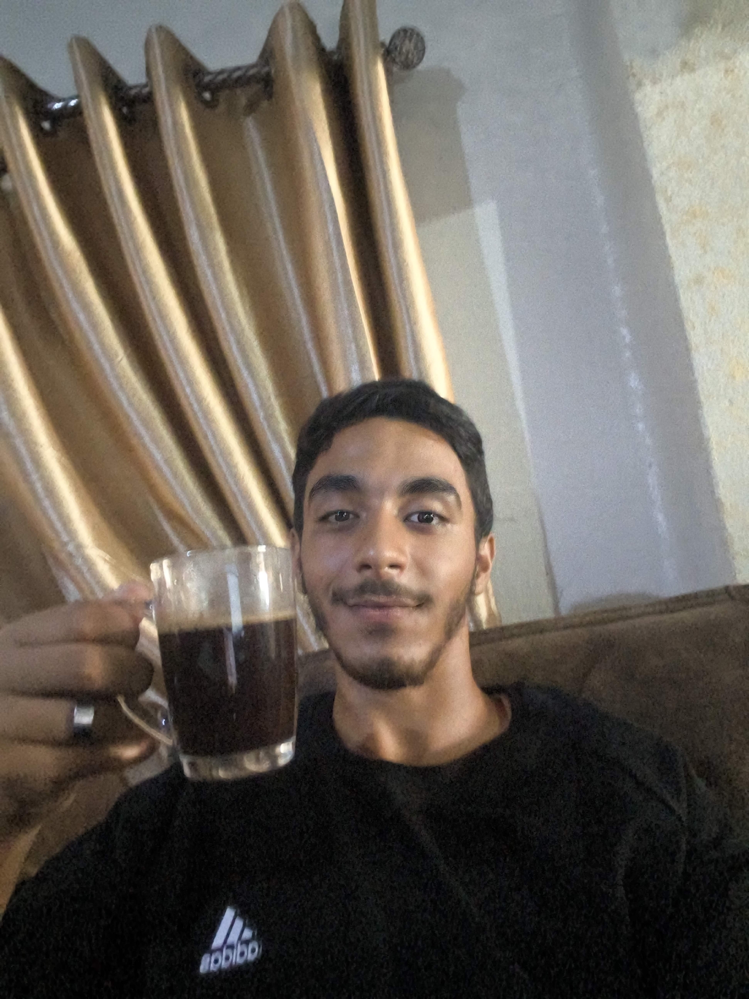 A young man is sitting indoors, holding a clear glass of dark liquid, possibly coffee, in his left hand. He is wearing a black textured sweater with a white Adidas logo visible on the chest. The background consists of golden curtains with vertical pleats, and a textured wall. The lighting suggests an indoor setting, possibly a living room. There are no visible indicators of a specific location within the city of Gaza, Palestinian Territories.