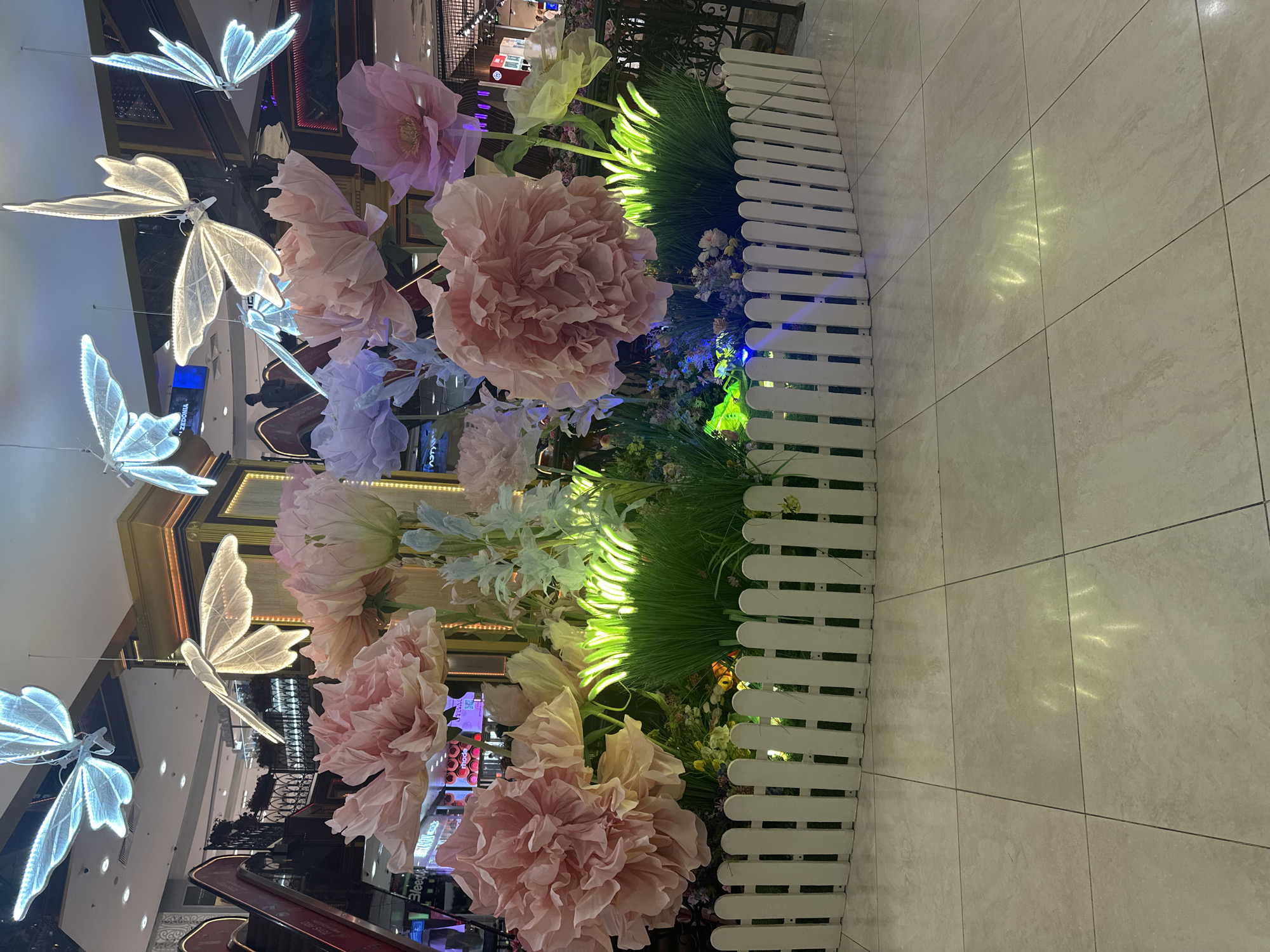 The image depicts an indoor decorative display, likely in a shopping mall. The central focus is a large, artificial floral arrangement featuring oversized pink and pastel-colored flowers resembling peonies or roses, interspersed with greenery that is illuminated from below with a vibrant green light. Surrounding this floral display is a short, white picket fence, curved to create a semi-circular enclosure. Above the flowers, several large, illuminated decorative butterflies, made of white and light blue light strips, are suspended from the ceiling. The background reveals a commercial interior with escalators, storefronts, and other shoppers. The floor is tiled in a light beige color with visible grout lines. While the scene is detailed, there is no specific information available to pinpoint the exact location within Moscow, Russia.