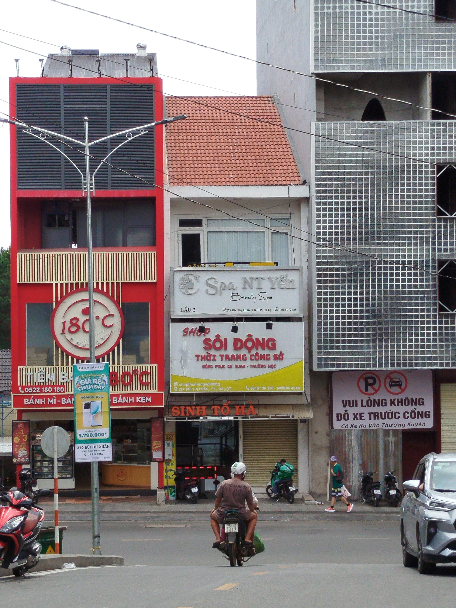 In Phường Xuân Hương - Đà Lạt, Vietnam, a street scene unfolds with a focus on commercial establishments and pedestrian activity. Three buildings of varying architectural styles dominate the foreground. The leftmost building is painted red and features a prominent circular logo with "180°C" and a heart symbol. Below this, signage indicates "BÁNH MÌ - BÁNH KEM," suggesting a food establishment. A streetlamp with a distinctive curved design stands tall in front of this building. To its right, a white building displays a sign for "Spa NT Yến Beauty Spa." Further to the right, a building clad in a grid of glass blocks stands, with a sign at its base reading "VUI LÒNG KHÔNG ĐỖ XE TRƯỚC CỔNG" (Please do not park in front of the gate), accompanied by a parking prohibition symbol.
On the street, several individuals are present. A man wearing a light brown t-shirt and white helmet rides a motorcycle, facing away from the camera. Another man in a green jacket rides a motorcycle to his left, also facing away. A third person, wearing a green t-shirt, blue shorts, and white shoes, walks on the sidewalk towards the right, carrying a white bag. Two motorcycles are parked on the sidewalk to the right of the glass-block building. In the background, trees are visible behind the red building, and a modern silver car is parked on the right side of the street. The overall environment suggests a busy urban setting with commercial activity and daily routines.