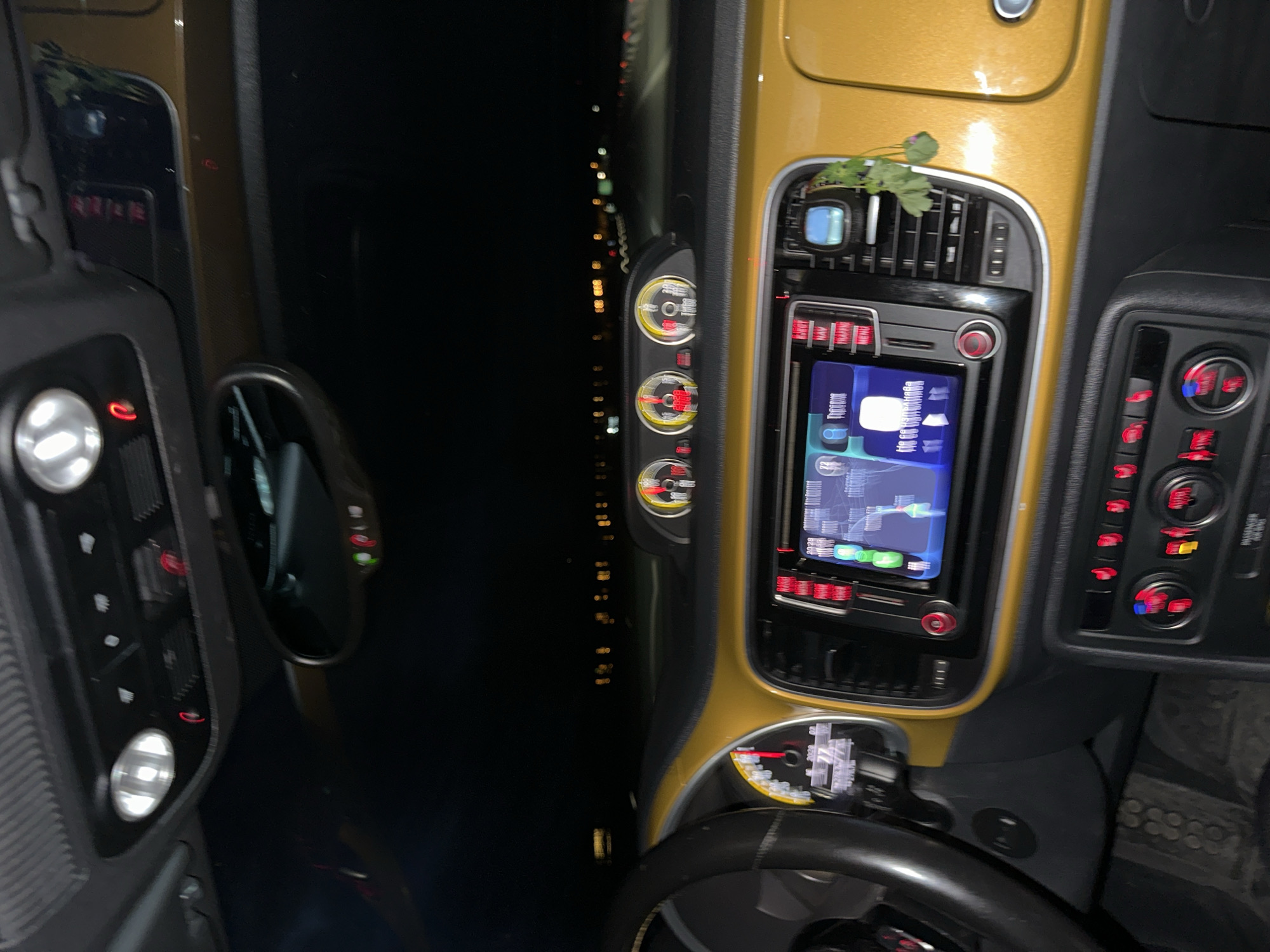 The image shows the interior of a vehicle, focusing on the dashboard and steering wheel. The dashboard has a gold finish, with a central console featuring a screen displaying a map or navigation system. There are also control panels with various buttons and dials. To the left of the dashboard is a side mirror, and beyond that, a dark expanse visible through the car window, with faint, blurred lights suggesting an urban or developed area at night. Considering the request for the location in Kato Ambelia, Greece, and the presence of blurred lights in the distance, it is suggested that the car is parked facing the city. There is no obvious indication of people or subjects other than the implied presence of a driver.