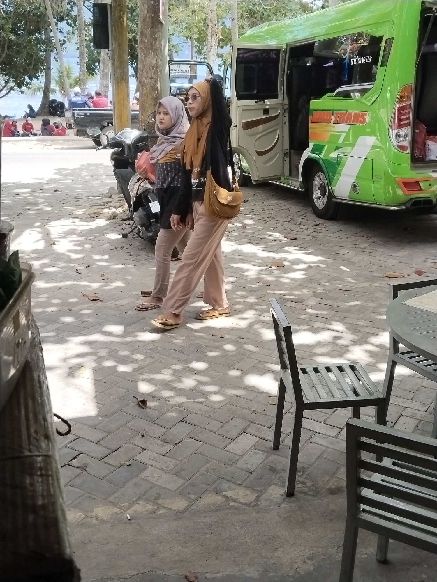 This image captures a street scene in Prigi, Indonesia. Two women, both wearing headscarves and modest clothing, are the central figures. The woman on the left has a light purple headscarf and is wearing a dark top with a patterned collar and light-colored pants. The woman on the right wears a light brown headscarf, a black jacket, and light-colored pants. She has a brown shoulder bag. They are standing on a brick-paved walkway.
Behind them, a green and white van with "BMD TRANS" written on its side is parked. The van's doors are open. To the left of the van, a motorcycle is visible. In the background, there are trees, a body of water that appears to be the sea, and a beach with several people sitting and standing. Some individuals are near a pickup truck. The ground is paved with rectangular bricks, and dappled sunlight creates shadows from the trees overhead. In the foreground, a metal chair and a table are partially visible, suggesting an outdoor seating area.