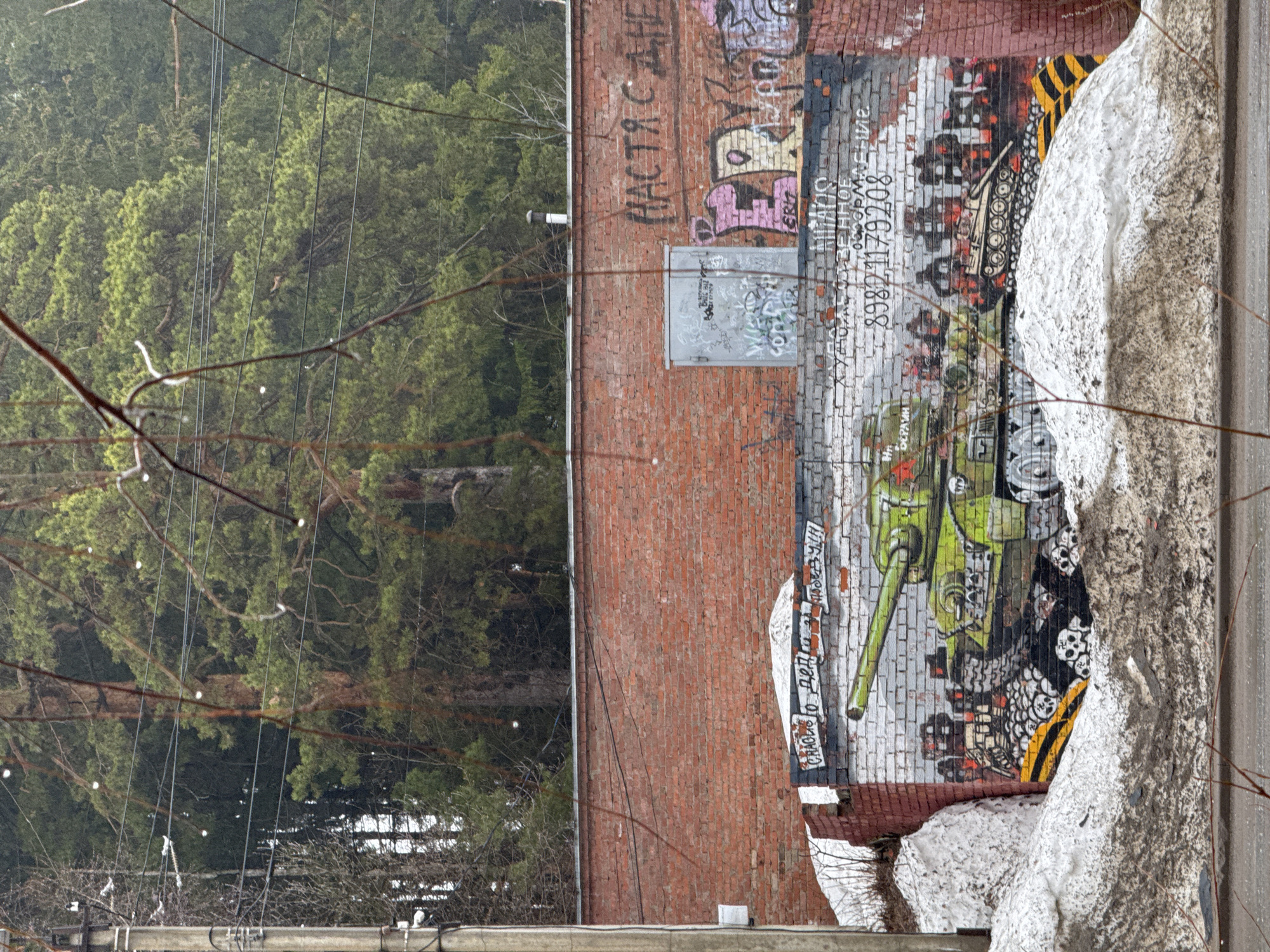 The image shows a brick building with graffiti in Votkinsk, Russia. The graffiti includes Russian script, a green tank, and the phone number 89821179208. The top of the wall is covered in old and recent graffiti. Next to the wall is a large pile of snow. In the background is a forest and telephone wires. The weather appears to be gloomy, possibly raining or snowing. Bare tree branches are in the foreground.
