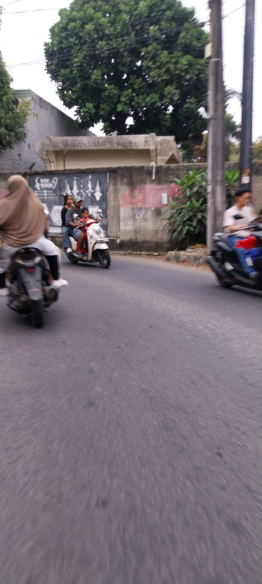 In the city of Kelapa Dua, Indonesia, a busy street scene unfolds, with several people riding motorbikes. On the left side of the frame, a person wearing a brown hijab rides a scooter. To their right, a white scooter carries three people: a man in a grey shirt and blue jeans, a woman in a black shirt, and a child in a red shirt. The man is driving, with the woman behind him and the child in front. Further to the right, another motorbike is partially visible, with a person wearing a white t-shirt and blue jeans riding it. The background features a low wall with a mural of koi fish, surrounded by buildings and lush green trees. The sky is overcast, suggesting it might be a cloudy day. The road appears to be asphalt, with some slight wear and tear.