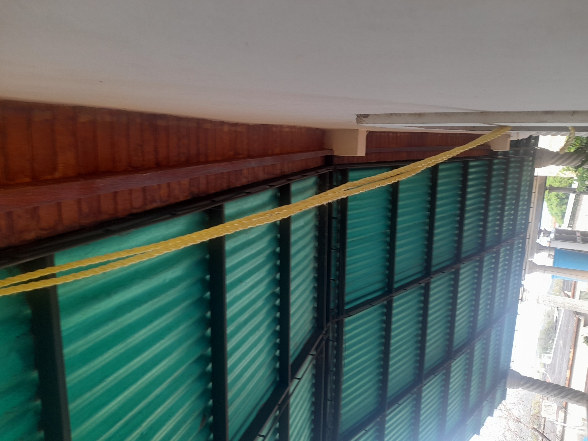 This is an upward angled, full shot of a covered outdoor area. The roof is made of corrugated green material supported by a black metal frame. On the right side, a wall painted white is visible. A thick yellow rope hangs down from the upper right of the frame, draping across the roof and the wall. The scene appears to be an outdoor patio or covered walkway.