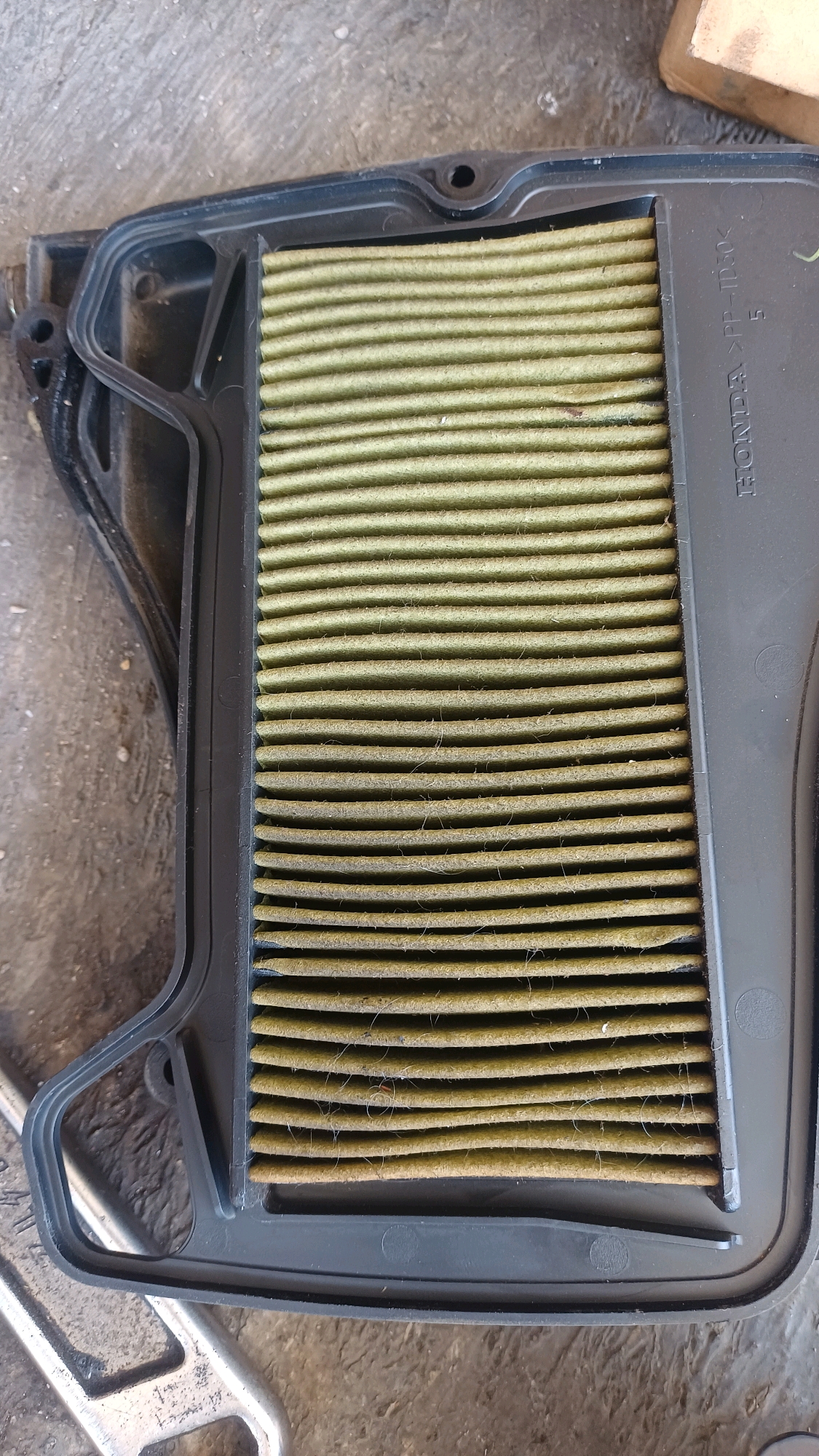 A top-down, medium shot captures a dirty, rectangular air filter for a vehicle, likely a motorcycle, resting on a grey concrete surface. The filter media is a light olive color with horizontal pleats, showing significant accumulation of dust and debris, indicating it needs replacement. It is housed in a black plastic frame, which is slightly elevated from the concrete by some unseen support. The frame has some embossed text that reads "HONDA >PP-TD30<". The background is the rough texture of the concrete. To the bottom left, a portion of a silver metal wrench is visible, suggesting the air filter is in the process of being removed or installed. The lighting is neutral, providing a clear view of the object and its surroundings. No people are present in the frame. The overall scene suggests a maintenance or repair setting, possibly a garage or outdoor workshop.