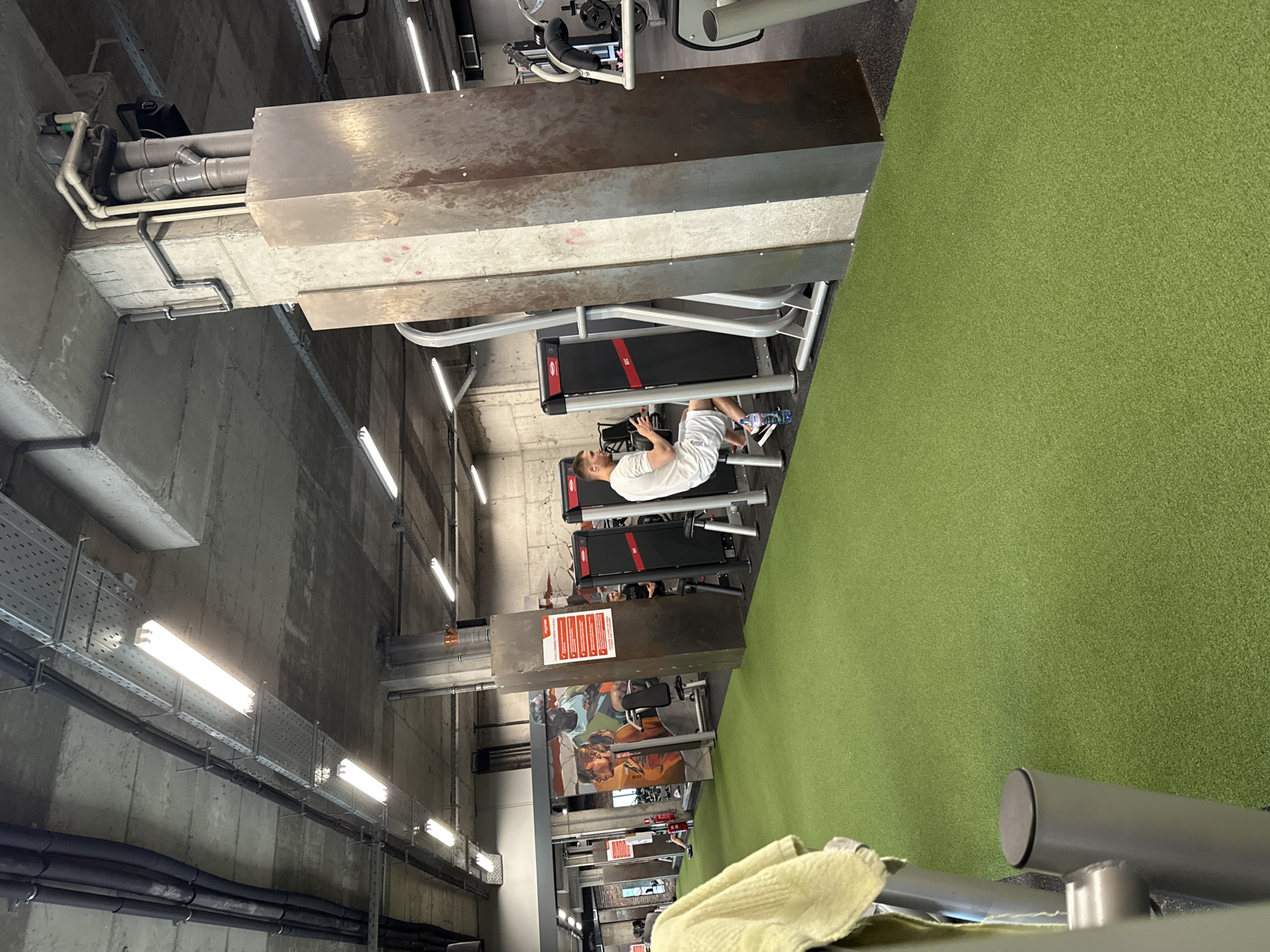 The image captures the interior of a modern gym or fitness facility in Sofia, Bulgaria, featuring a distinct industrial aesthetic. Exposed concrete walls and ceilings are prominent on the left and top, showcasing an array of utility infrastructure including grey and white pipes, black insulated tubing, and metal cable trays fitted with fluorescent strip lights.
A large section of the floor on the right is covered with vibrant green artificial turf. Several pieces of gym equipment are arranged along the divide between the concrete and turf areas. In the mid-ground, a man dressed in a white t-shirt, white shorts, and sneakers is seated on a leg press machine, with a blue water bottle near his feet. Behind him, two treadmills with red accents are visible.
Large, rectangular metal structures, possibly ventilation ducts or utility enclosures, span overhead. These structures have a weathered, rusted appearance. One such structure features a red sign with multiple lines of white text, likely providing warnings or instructions. Further into the background, a colorful mural or large poster depicting human figures engaged in what appears to be athletic activity is mounted on a wall. In the immediate foreground, a light-colored towel rests on a piece of gym equipment. The setting is indoors, illuminated by artificial lighting.