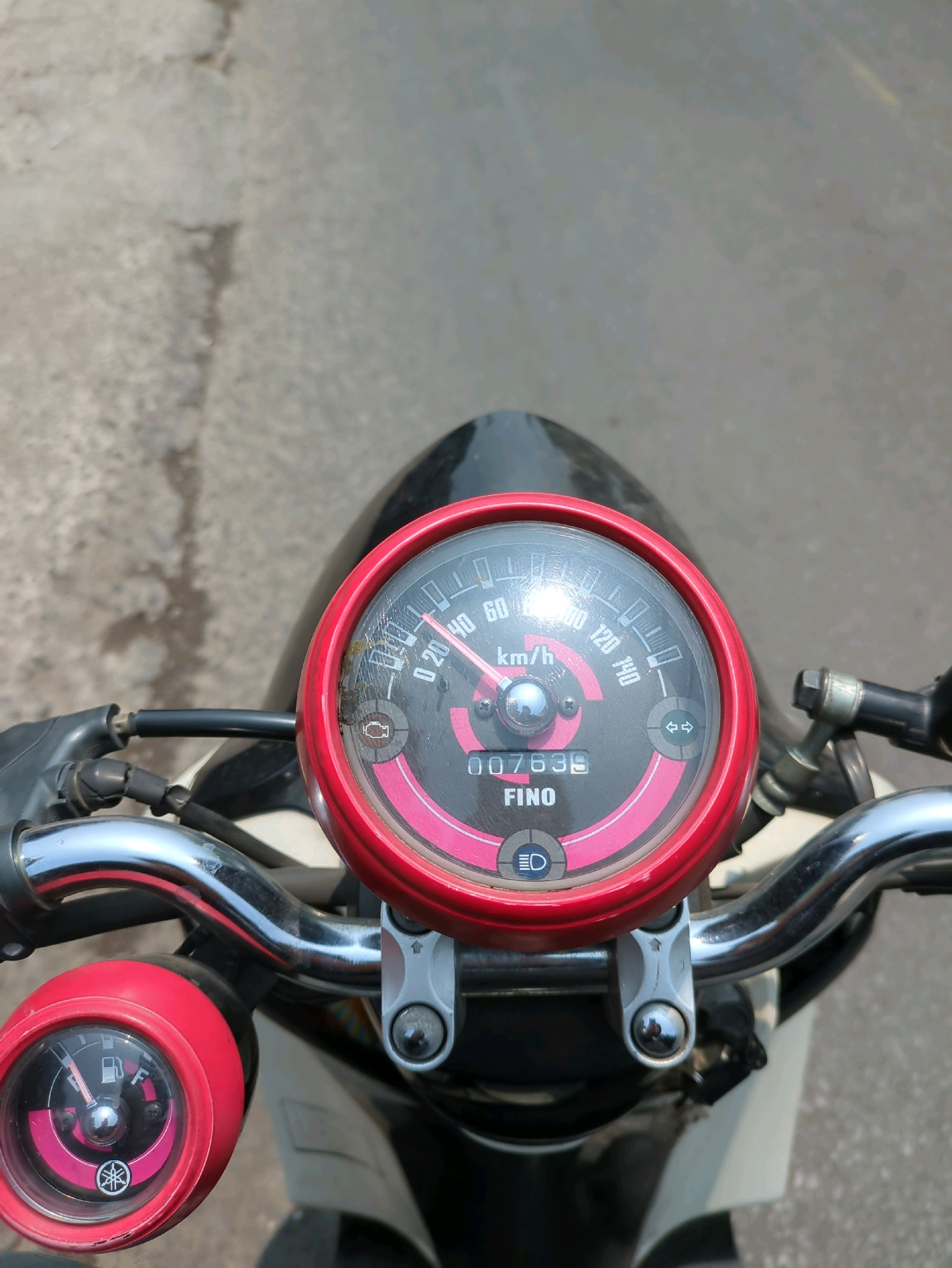 The image shows a close-up view of a Yamaha Fino scooter's handlebars and instrument cluster. The scooter is parked on an asphalt road, which appears to be in Gumpang, Indonesia, based on the contextual information provided. The instrument cluster is circular with a red bezel and features a speedometer with markings up to 140 km/h, a digital odometer displaying "007635," and the "FINO" model name. A fuel gauge is visible on the left side of the handlebars. The environment is outdoors, with a neutral-toned, textured asphalt surface filling the background. There are no people visible in the image.