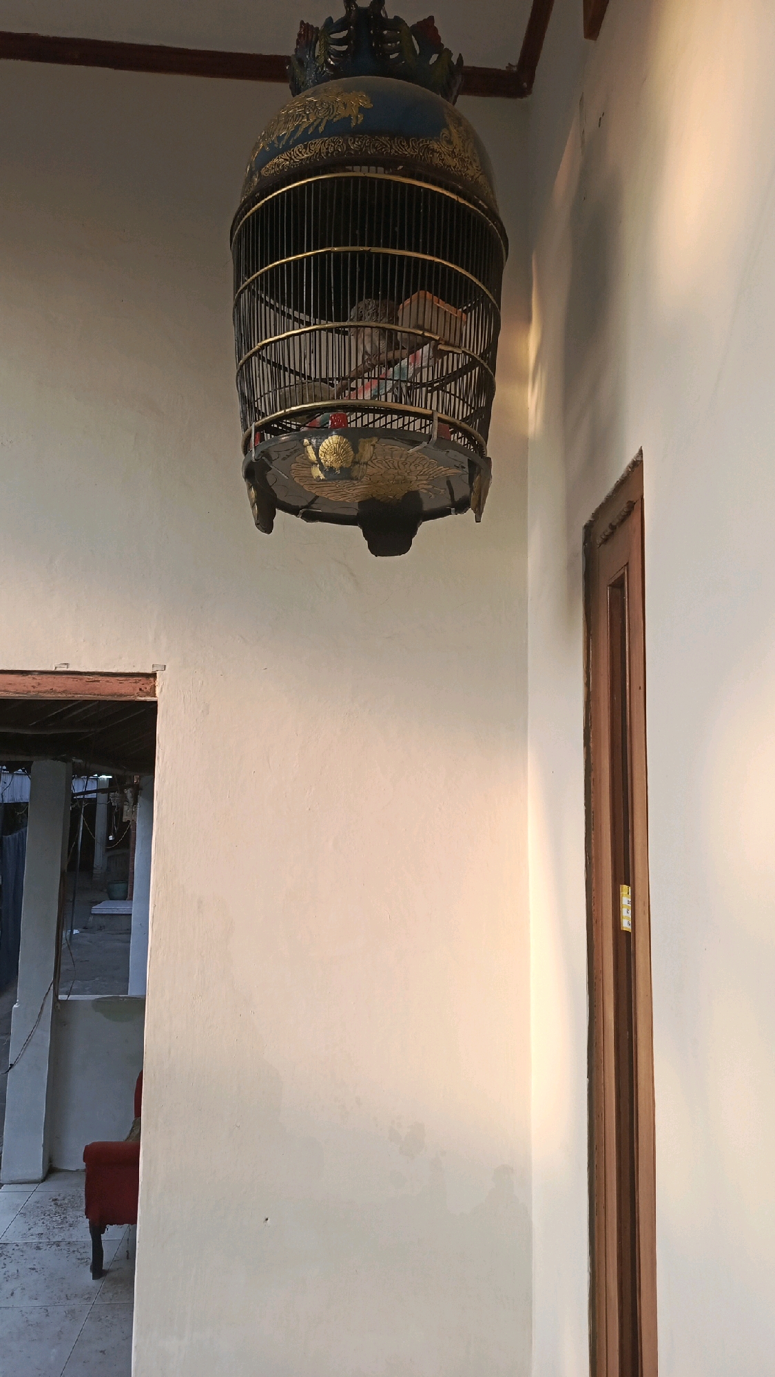 A vintage, ornate birdcage hangs from the ceiling of an interior space. The cage is primarily made of dark metal bars with a decorative, possibly gilded, base and lid. Inside the cage, a small bird can be seen. The walls of the room are painted a light color and appear textured. Sunlight is casting a warm glow on the right wall, creating a gradient of light and shadow. To the left of the frame, an open doorway reveals a glimpse of another area with a red chair. A wooden doorframe is visible on the right side of the image. The scene is located in Pekauman Kulon, Indonesia.