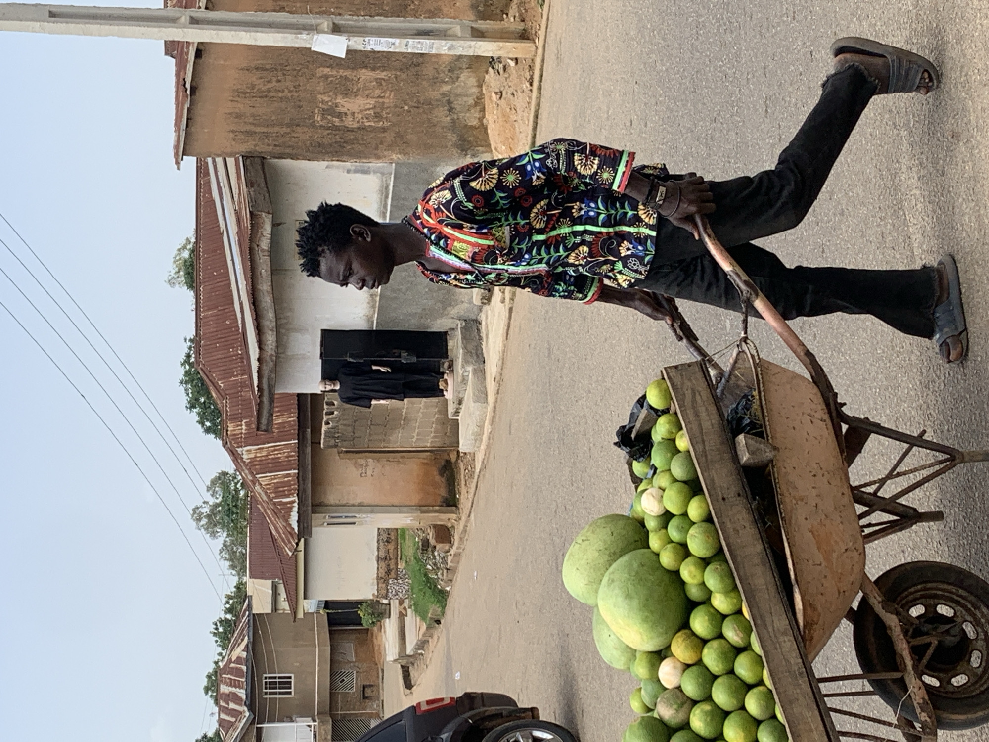 In the city of Jos, Nigeria, a young Black male is pushing a wheelbarrow filled with green fruits, likely citrus, and two large, round, light green fruits, possibly watermelons, on a paved road. He is wearing a black shirt with a colorful, floral pattern in red, yellow, blue, and green, black pants, and dark sandals. His hair is short and textured. The wheelbarrow is made of metal and wood. In the background, there are residential buildings with corrugated metal roofs and concrete walls, some appearing weathered. A dark-colored SUV is parked on the left side of the road. Two mannequins in black clothing stand in front of a building on the right. The sky is overcast. The scene depicts a street vendor in an urban Nigerian environment.