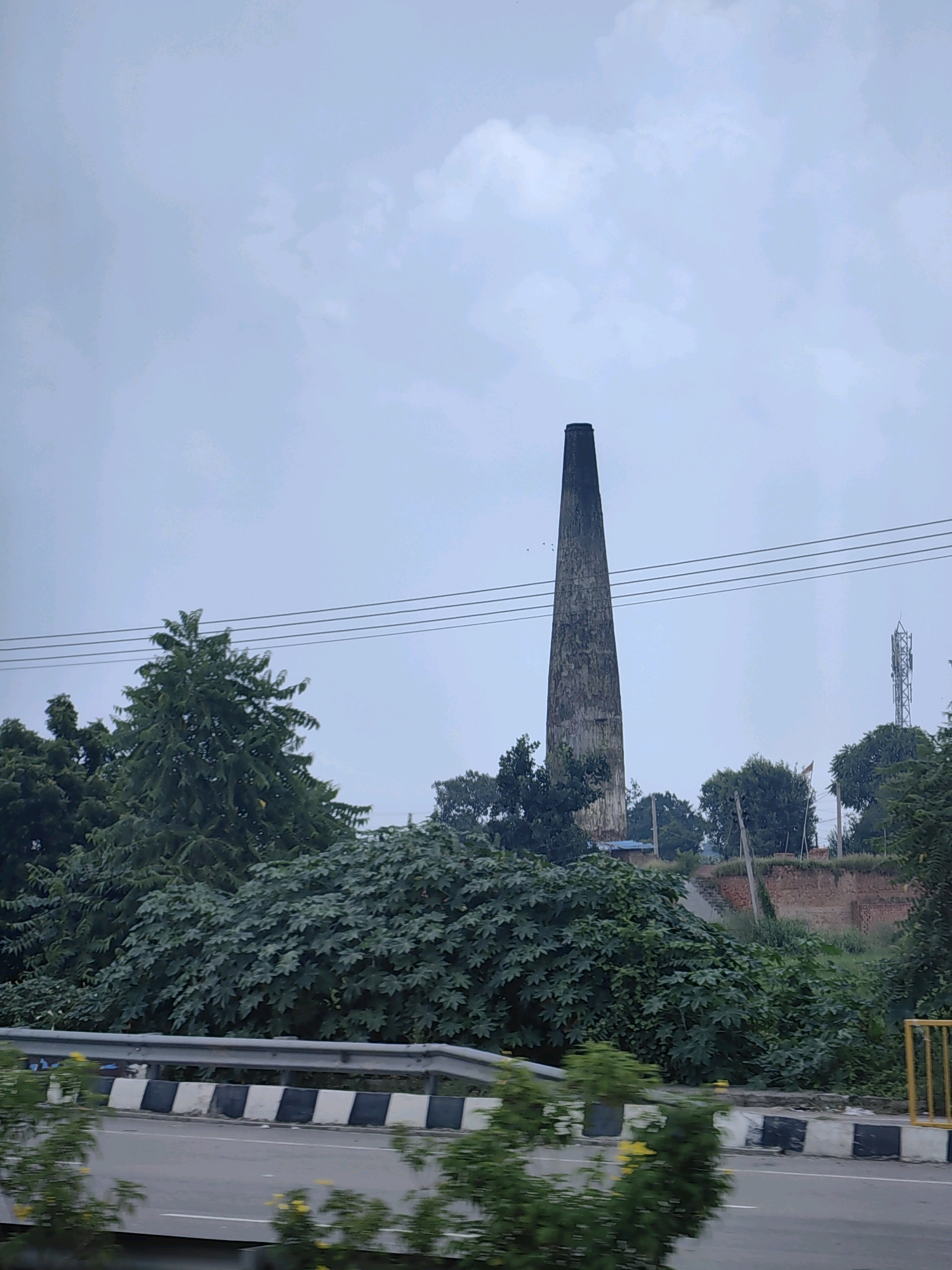 The image depicts a tall, conical brick chimney dominating the center of the frame, against a cloudy, overcast sky. The chimney appears weathered and textured, rising from a dense cluster of dark green foliage. In the mid-ground, a red brick wall is visible, partially obscured by the vegetation and a small cell tower stands to the right of the chimney. A paved road with a guardrail and black and white striped barriers runs along the bottom of the image, suggesting it is a roadside view. The overall environment appears to be rural or semi-industrial, with abundant plant life. The location is identified as Passina Kalan, India.
