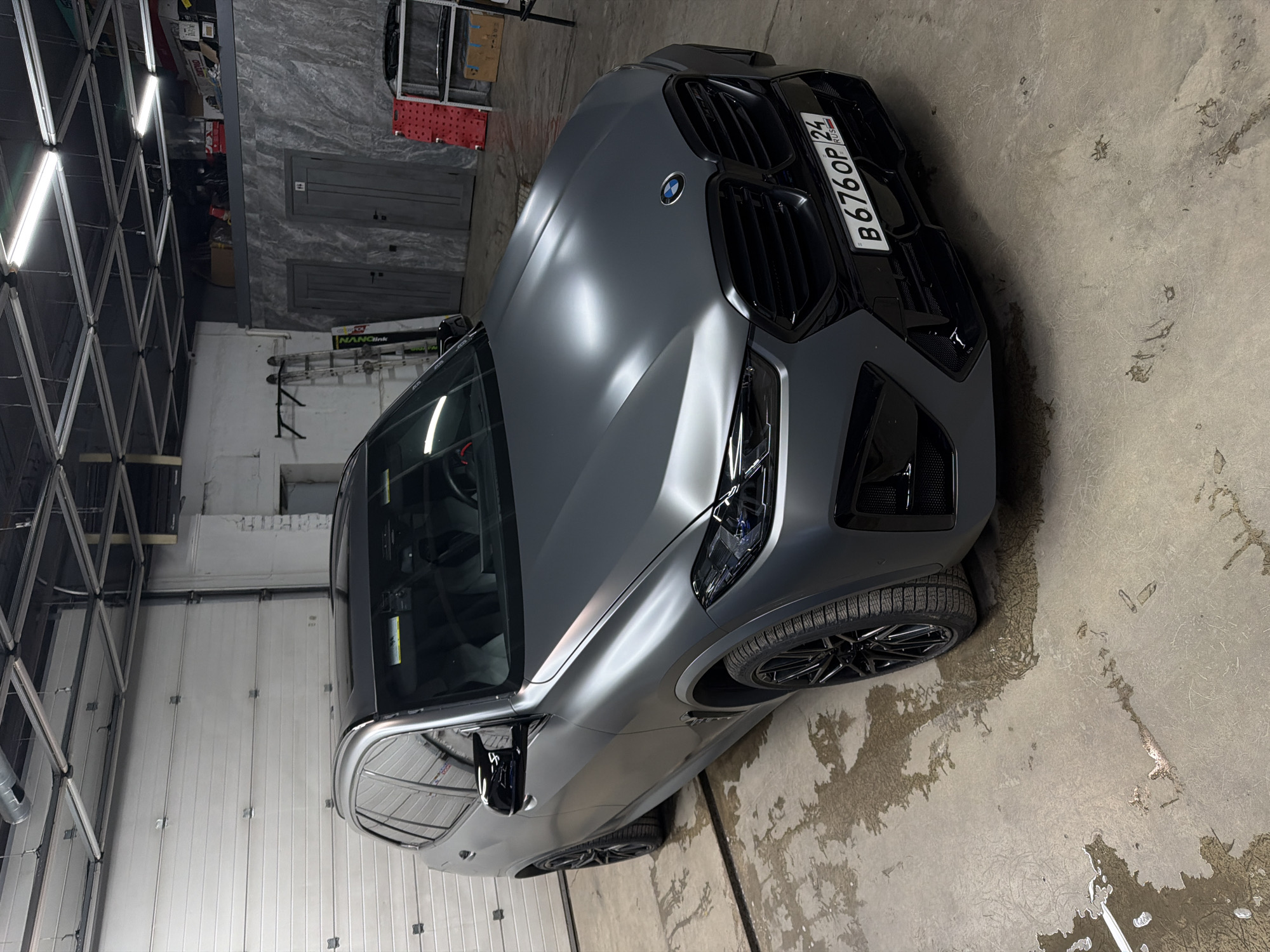 The image shows a gray BMW SUV parked in an indoor garage. The car's license plate reads "B 676 OP 24", indicating registration in the Krasnoyarsk region of Russia. The garage appears to be a vehicle maintenance or storage facility with tools, equipment, and a multi-panel garage door visible in the background. The floor is concrete with water marks and patterns.