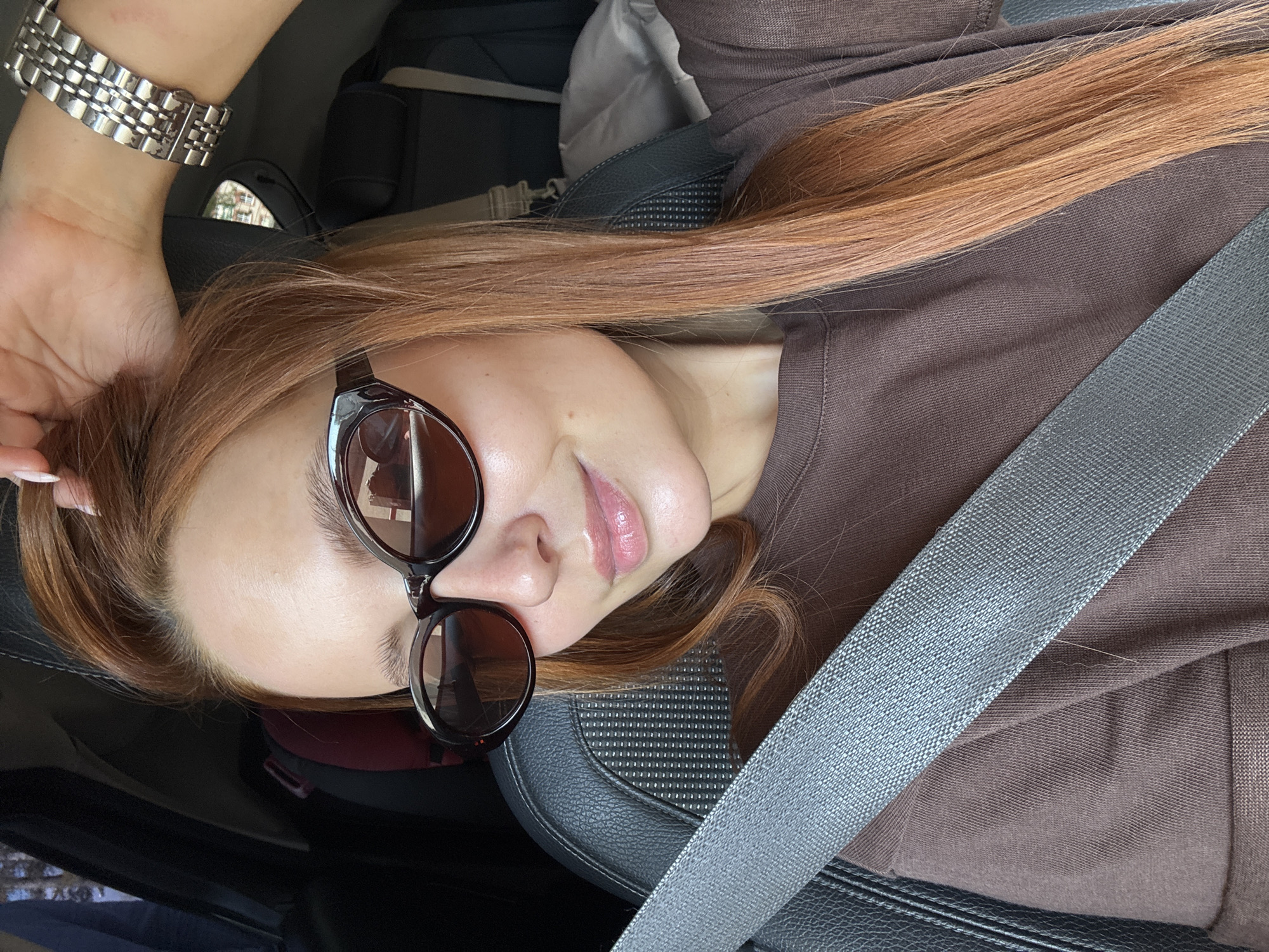A person wearing sunglasses is in the driver's seat of a car, with the seatbelt fastened. The person has reddish-blonde hair, light skin, and is wearing a brown shirt. Their left hand is raised, with a silver watch visible. The interior of the car is black and gray. The image location is unknown.