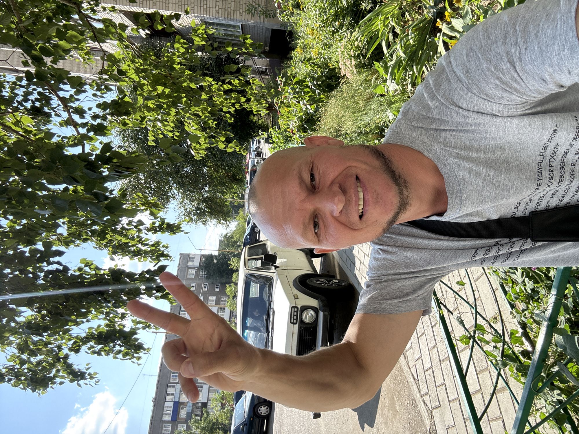 The image shows a man taking a selfie outdoors on a sunny day. He is bald, with a beard, and is wearing a grey t-shirt. He is holding up his right hand in a peace sign. Behind him, a white car is parked on the street. The background shows apartment buildings, trees, and a garden with lush green foliage. The man appears to be on a sidewalk next to a green fence. The location is identified as Oskemen, Kazakhstan.