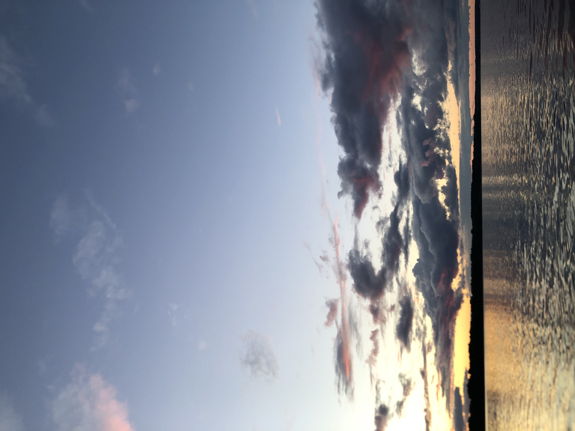 This image depicts a tranquil sunset over a body of water, with a silhouetted treeline forming the horizon. The sky is a gradient of blues and purples, transitioning to warm oranges and yellows near the horizon, where scattered clouds are tinged with pink and grey. The water reflects the colors of the sky and the faint lights of the setting sun, creating shimmering patterns on its surface. The overall atmosphere is serene and picturesque, suggesting a peaceful evening in a natural setting. The location is identified as Svirstroy, Russia.