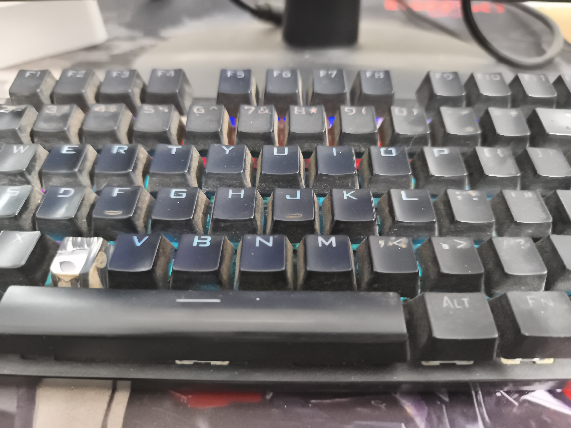 A close-up, eye-level shot shows a section of a black keyboard with backlit keys. The keys are dusty, indicating they are in use. The top row of keys displays function keys like F1 through F11. Below this are number keys and symbol keys, followed by the QWERTY alphabet keys. The image captures specific letters such as W, E, R, T, Y, U, I, O, P, A, S, D, F, G, H, J, K, L, Z, X, C, V, B, N, M, and modifier keys like ALT and FN. A unique keycap with a cartoon character design is visible on the left side of the spacebar. The keyboard is positioned on a desk or surface with a patterned mousepad underneath. The background is out of focus, showing a white object and a black cable. The lighting is ambient, with the colorful backlighting of the keyboard keys being a prominent feature. There is no indication of the city of Kutoarjo, Indonesia.