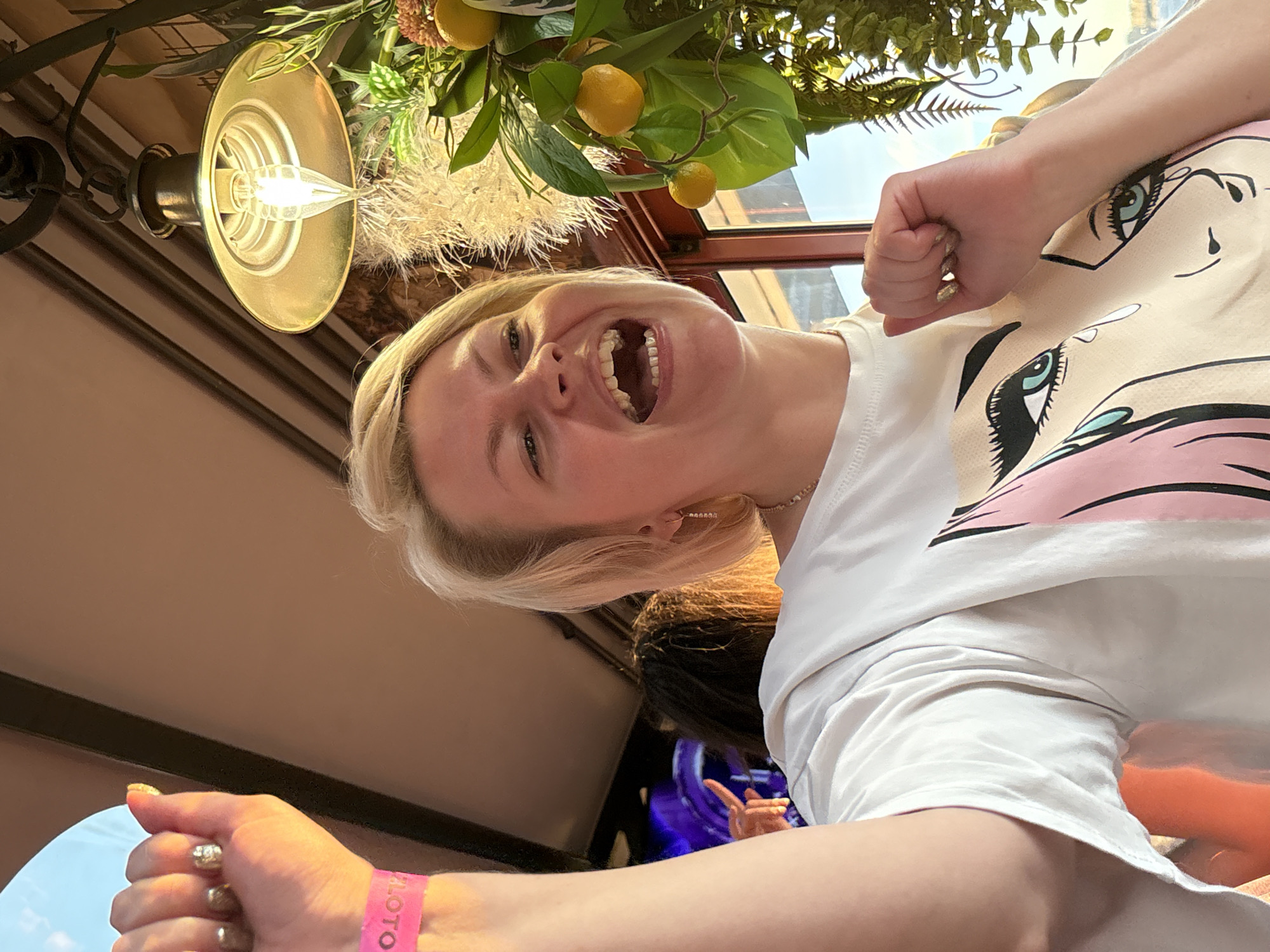 A woman with blonde hair is captured in a close-up, low-angle shot, laughing with her mouth wide open. She is wearing a white t-shirt with a graphic print of a crying cartoon face. On her left wrist, a bright pink wristband with the word "LOTO" is visible. She is raising her left arm with her fist clenched. In the background, out of focus, other people are visible, and a decorative plant with yellow fruits hangs from a lamp. The lighting suggests it is either indoors or evening outdoors. The scene appears to be in Saint Petersburg, Russia, possibly at a restaurant or event venue, given the decorative elements and the presence of a wristband which could indicate entry to a paid event.