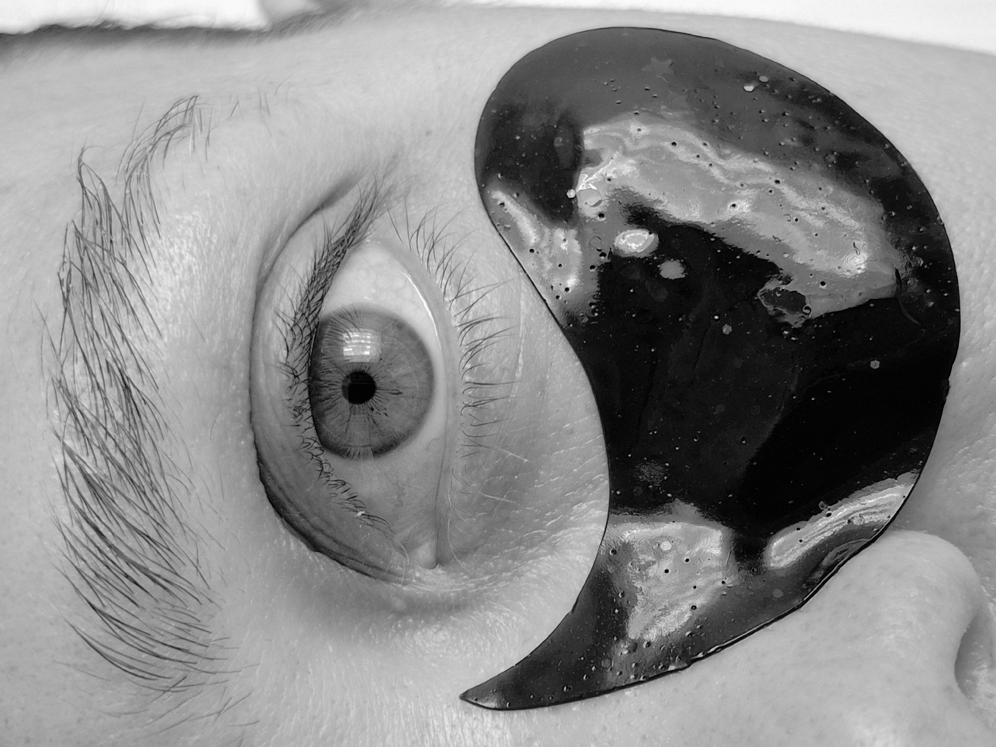 A close-up, black and white image shows a human eye with a dark, crescent-shaped object (possibly a cosmetic eye patch) placed beneath it. The eye is open and gazing directly ahead. Visible features include the eyebrow, eyelashes, and iris. The skin surrounding the eye is pale. The eye patch appears glossy with small imperfections. The location of the image is not discernable.