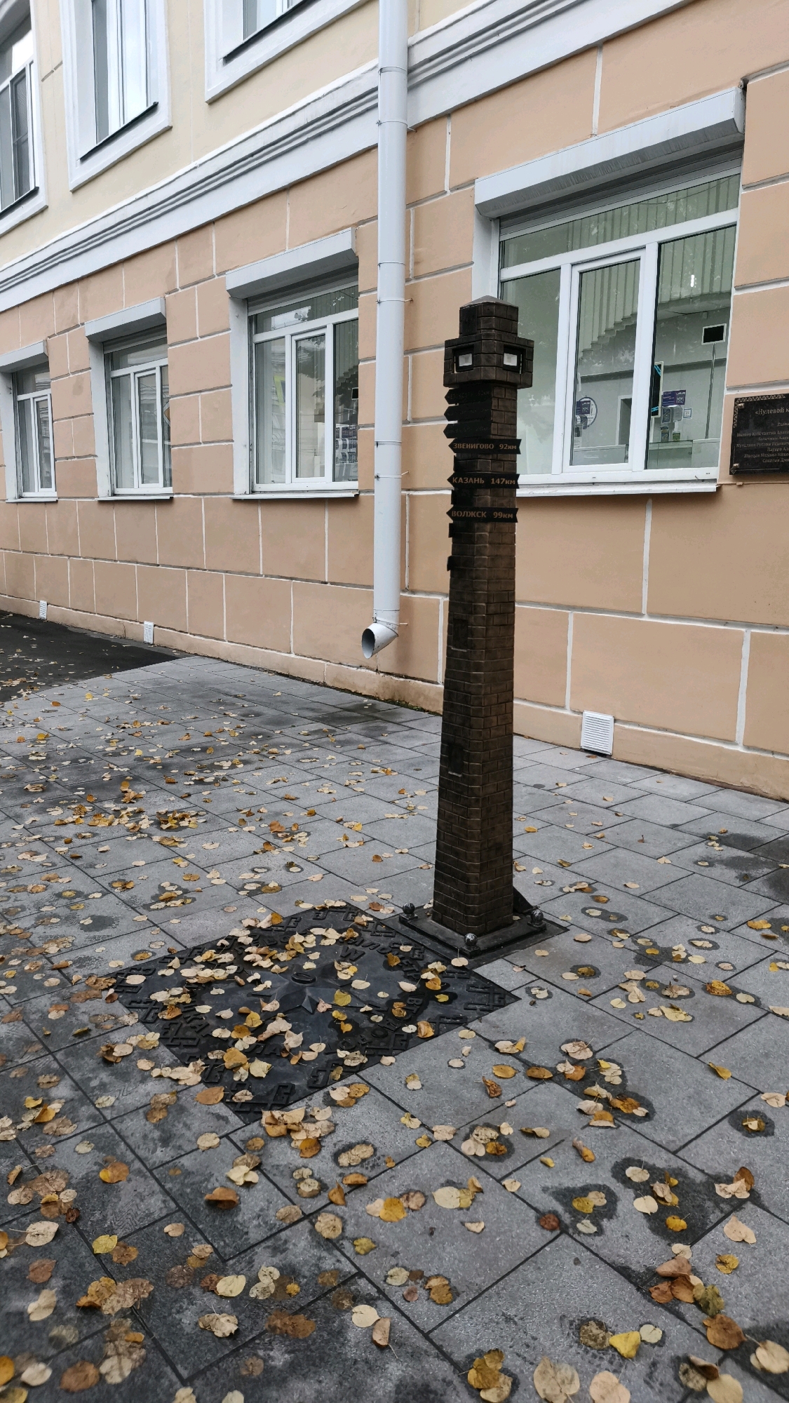 The image captures an outdoor scene in Yoshkar-Ola, Russia, featuring a brick monument with directional signs. The monument is situated on a paved walkway, which is partially covered with fallen yellow and brown leaves. Behind the monument is the facade of a building with light peach-colored walls and several windows. A white downspout runs vertically along the building's exterior, to the left of the monument. The monument itself is a dark, brick-like structure with protruding signs pointing in different directions, indicating distances to various locations. Some of these signs are legible, showing place names like "ЗВЕНИГОВО 92км" (Zvenigovo 92km), "КАЗАНЬ 147км" (Kazan 147km), and "ВОЛЖСК 99км" (Volzhsk 99km). In front of the monument, on the paved ground, is a square manhole cover with an embossed star-like design. The overall impression is a somewhat somber autumn day. There are no people or animals visible in the scene.