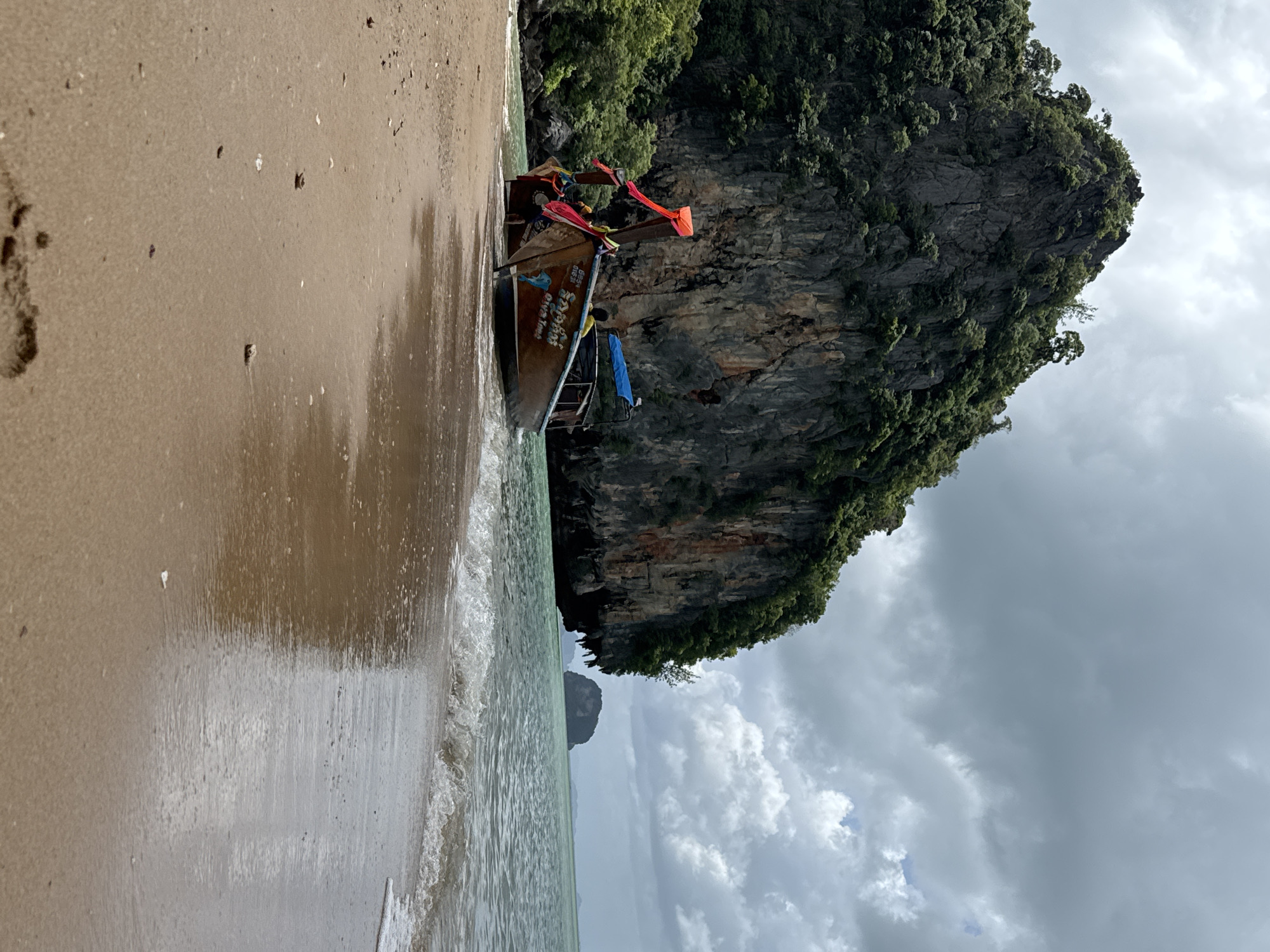 In Khlong Khian, Thailand, a longtail boat is beached on a sandy shore, its bow pointed towards the water. The boat is made of wood and has a blue canopy over the passenger area. It is adorned with colorful flags and ribbons. Behind the boat, a sheer, tree-covered limestone cliff rises dramatically from the water. The sea is calm with gentle waves lapping at the shore. The sky is overcast with dramatic clouds, casting a diffused light over the scene. In the distance, a smaller rocky island is visible. The foreground shows a sandy beach with scattered shells and debris.
