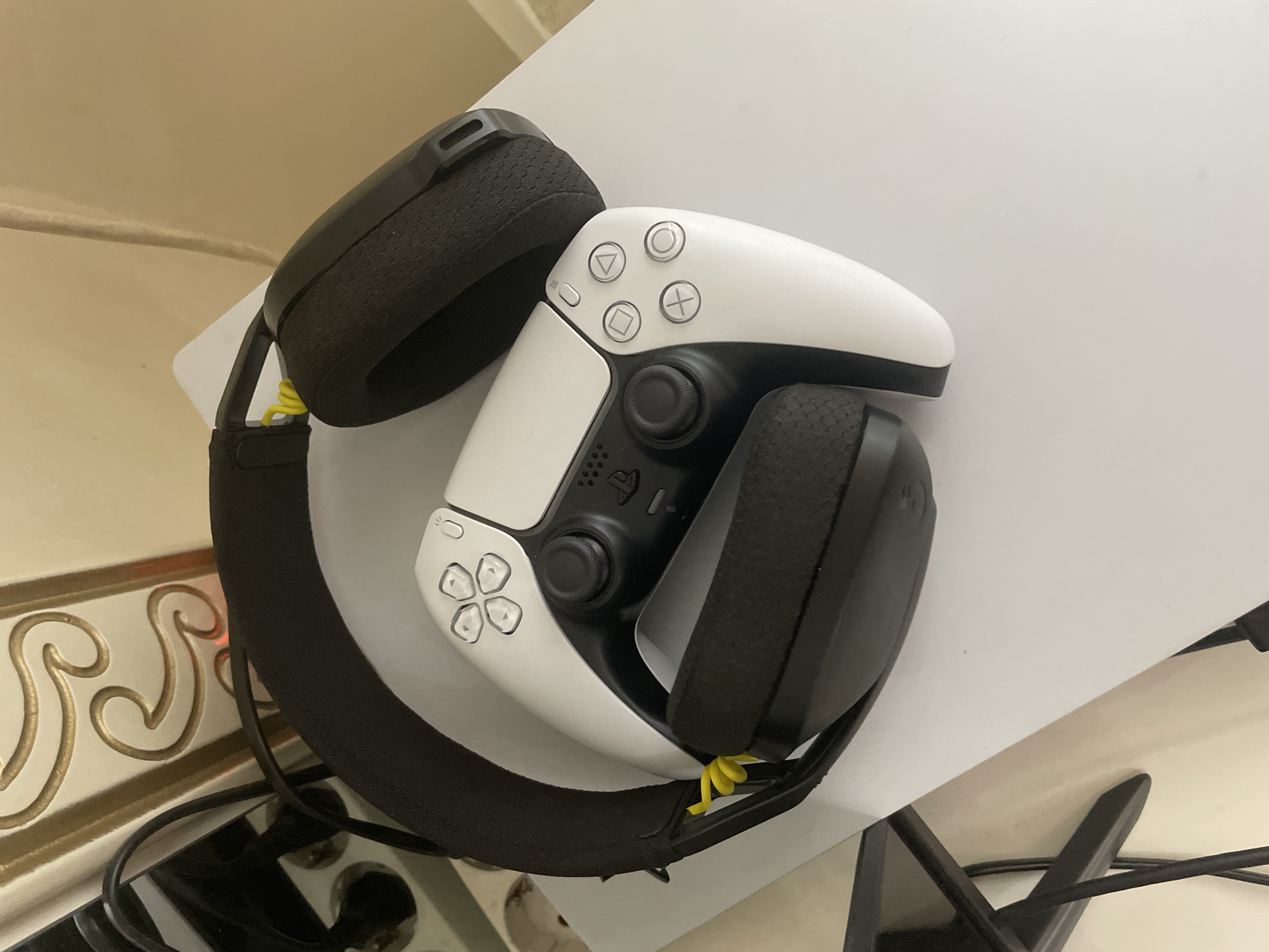 The image displays gaming accessories in an indoor setting. The primary subjects are a white Sony PlayStation 5 DualSense wireless controller and a black gaming headset.
The PlayStation 5 controller is positioned centrally, resting on the earcups of the headset. It is predominantly white with a black central panel, black analog sticks, and black trigger buttons. The iconic PlayStation face buttons – circle, cross, square, and triangle – are visible, appearing translucent with white outlines. A small PlayStation logo is etched into the central button below the analog sticks.
The gaming headset is black with substantial cushioned earcups and a padded headband. A distinctive bright yellow coiled elastic band or cable tie is visible securing a part of the headset near one of the earcups. The entire setup rests on a clean, flat white surface, which could be a console itself or a dedicated stand.
The background includes a decorative element to the lower left, featuring an intricate, raised golden wavy pattern against a light beige or cream-colored surface, possibly part of furniture or wall decor. In the bottom right corner, a black, angular structure, likely a furniture leg or stand, is visible along with some black cables. The lighting is even, suggesting an indoor environment. No activity is depicted; the items appear to be stored or temporarily set aside. There is no discernible text beyond the standard PlayStation button symbols.
The image was captured in Ihtiman, Bulgaria.