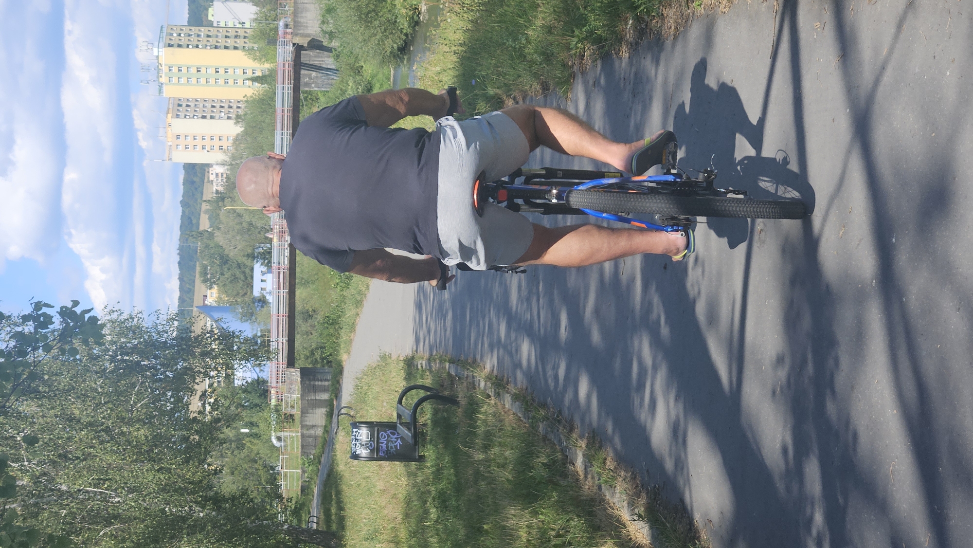 The image shows a bald man riding a bicycle on a paved path in the city of Prešov, Slovakia. The man is wearing a dark gray t-shirt and light gray shorts, and flip-flops on his feet. He is viewed from behind. The path runs alongside a grassy area with trees and a river or canal. In the background, there are buildings and a bridge, characteristic of an urban environment. The sky is blue with some clouds. The scene appears to be a sunny day. The path is shaded in places by the trees. There is a bench with graffiti on it to the left of the path.