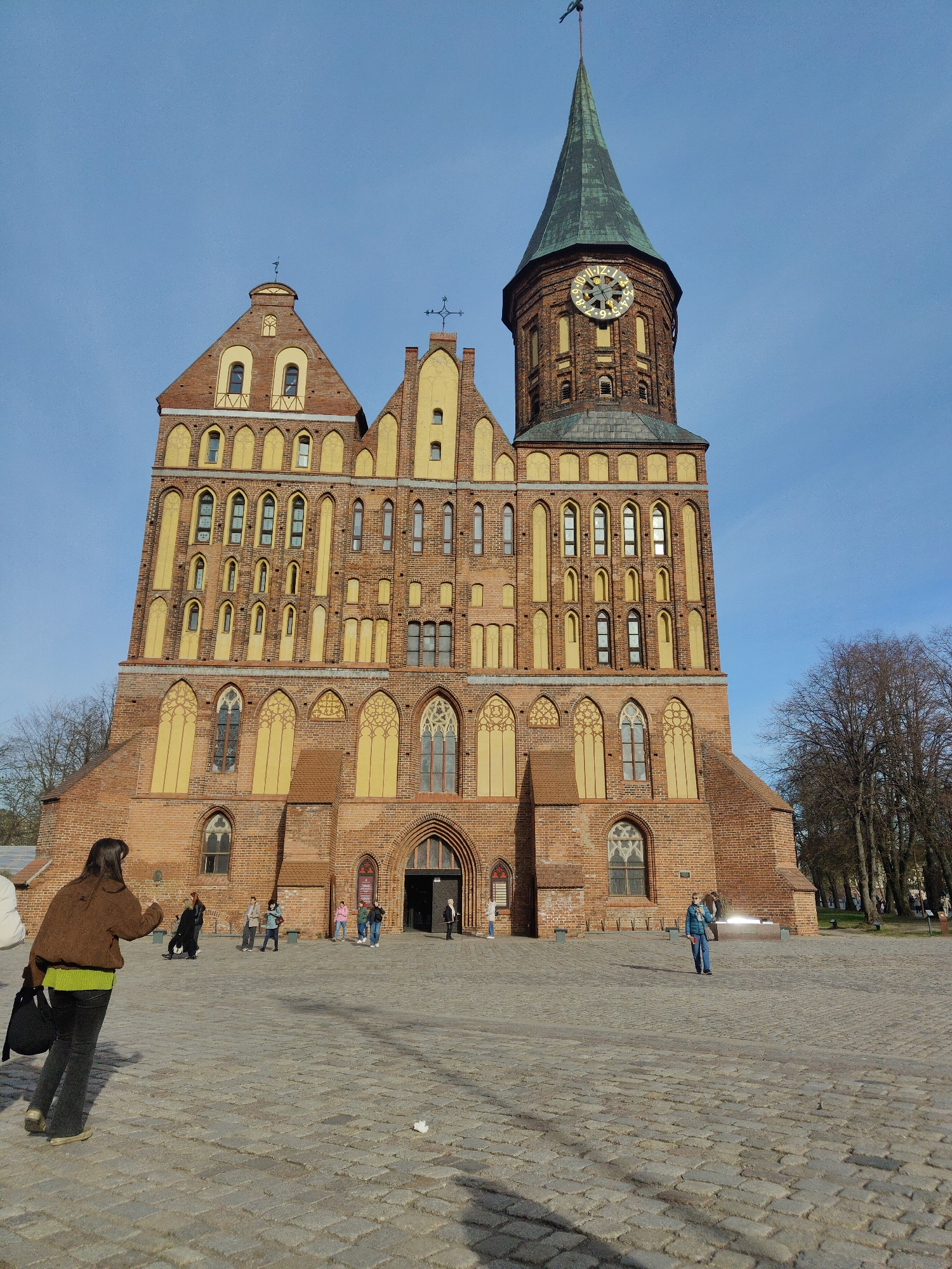 A large, imposing brick building, likely a cathedral or church, dominates the center of the image under a clear blue sky. The building features red brickwork contrasted with yellow decorative elements around its numerous arched windows and gables. On the right side, a tall, octagonal clock tower with a dark green spired roof stands out. The clock face is round, gold-colored, and shows radial lines indicating hours, with some numerals visible.
In the foreground, a woman with dark hair, wearing a brown jacket and a lime green top, walks away from the camera across a cobblestone square, carrying a black bag. Several other individuals are scattered across the square and near the building's main arched entrance, some appearing to be tourists or visitors. On the far right, a woman in a blue jacket stands in the square. Bare trees line the right edge of the square, suggesting a late autumn or early spring season. The bright daylight creates distinct shadows on the ground and building, indicating a sunny day, likely late morning or early afternoon. The overall scene depicts a historical site being visited by people. No clearly legible text is visible apart from the general design on the clock face.