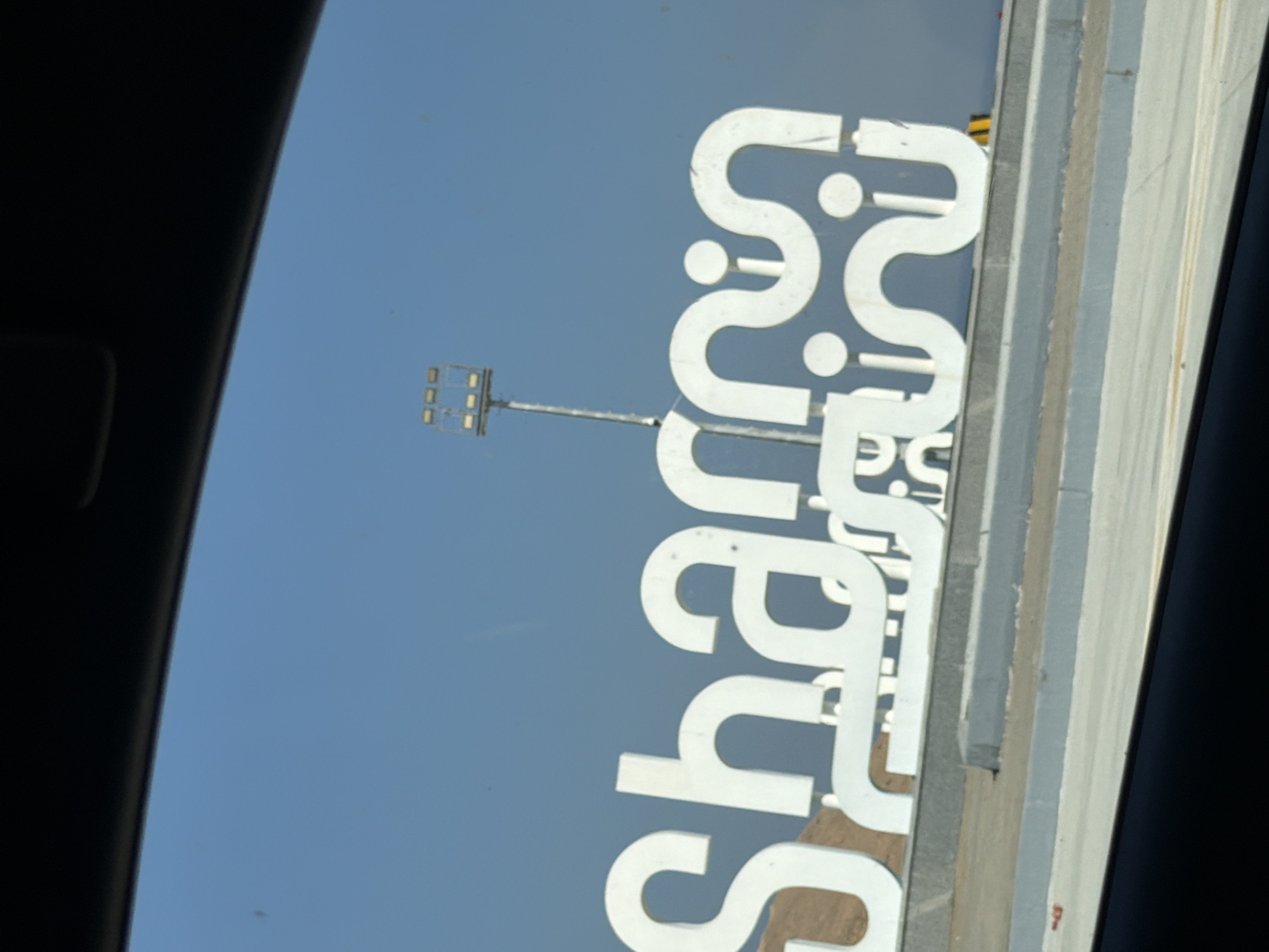A large, white, stylized sign with Arabic lettering is positioned on a slight incline under a clear blue sky. The sign, which appears to spell "Sharm," is illuminated by a tall pole with multiple light fixtures on top, visible behind the right side of the sign. The lettering is bold and abstract, with curves and dots. The sign is mounted on a concrete base with grey and brown accents. In the foreground, a dark area, possibly the interior of a vehicle, frames the bottom of the image. The overall scene suggests an outdoor setting in Sharm El Sheikh, Egypt, likely a public area or entrance.