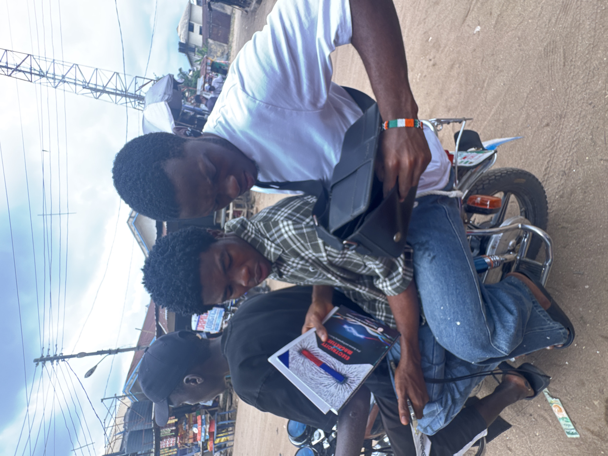 Three young men are situated on a parked motorcycle within the urban setting of Ihiagwa, Nigeria. One individual, wearing a white shirt and blue jeans, appears to be retrieving something from a black shoulder bag. Another man in a plaid shirt is seated behind him, holding a textbook titled "Electricity Machine." A third individual, wearing a black shirt and cap, is partially visible in the lower left corner. The environment includes buildings, power lines, and street signs indicative of a populated area.