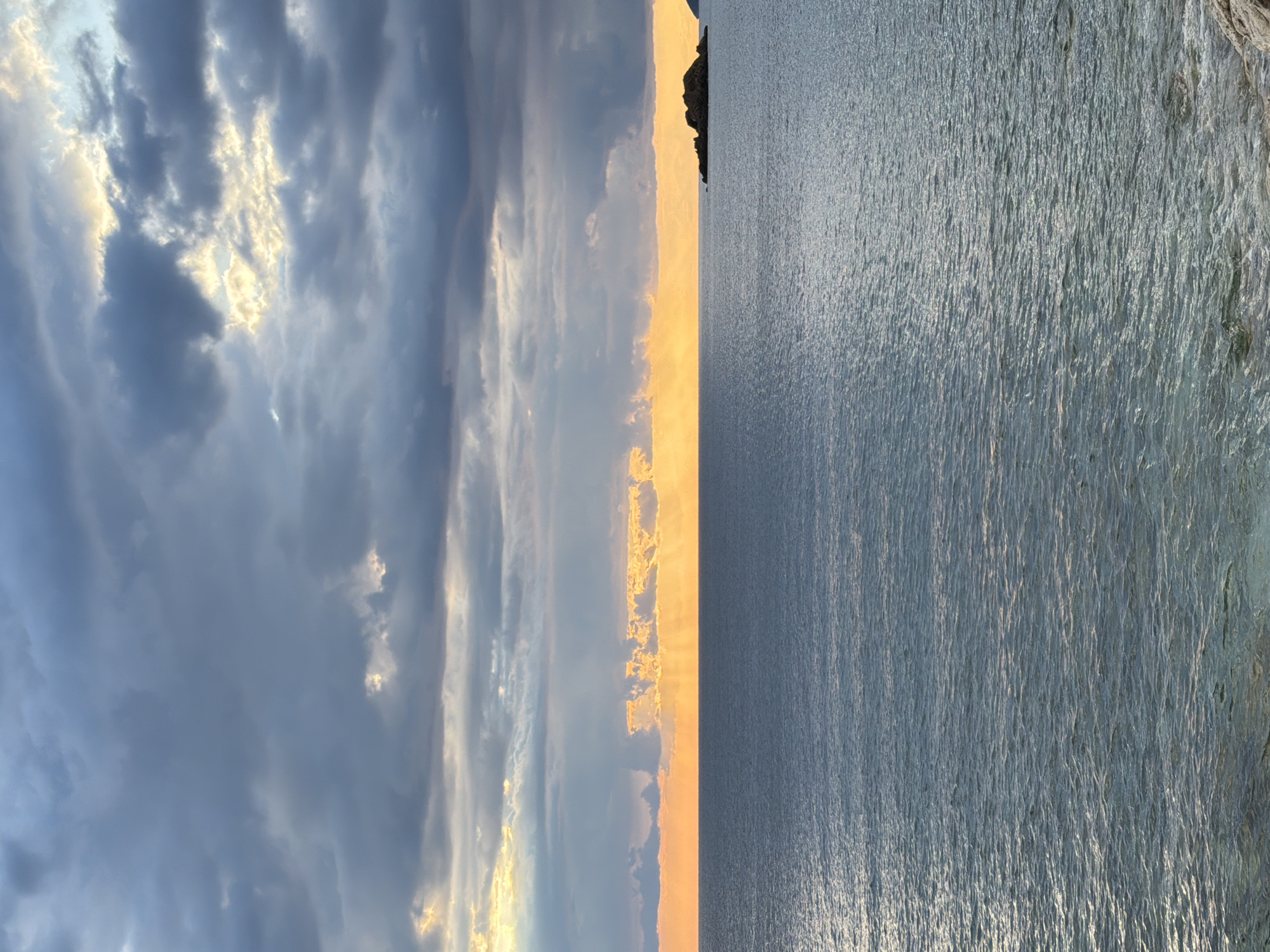 The image shows a vertical view of a seascape at sunset in Perazića Do, Montenegro. The left side is dominated by a sky filled with dramatic, gray, layered clouds illuminated by golden sunlight. A narrow strip of the horizon is awash in the golden glow of the setting sun, creating a vibrant line separating the sky from the sea. The right side of the image displays the calm, textured surface of the water, reflecting the muted colors of the sky and sun. In the distance, a dark, rocky promontory juts into the sea. The overall impression is one of tranquility and natural beauty.