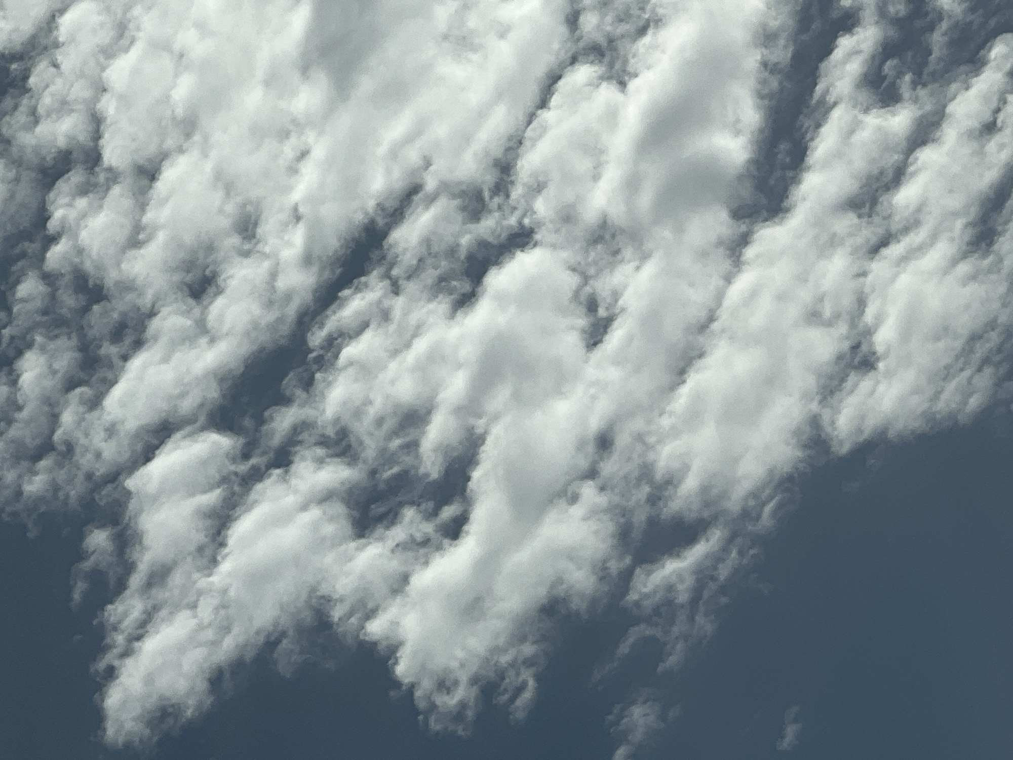 The image shows a close-up view of a sky filled with white, fluffy clouds. The clouds are arranged in horizontal bands with spaces of blue sky visible between them. The lighting suggests it is daytime, with the sun illuminating the tops of the clouds, creating shadows on their undersides. The image does not contain any identifiable landmarks or elements that would allow for the determination of a specific location in Warsaw, Poland, or any other city. The scene is solely focused on the atmospheric conditions of the sky.