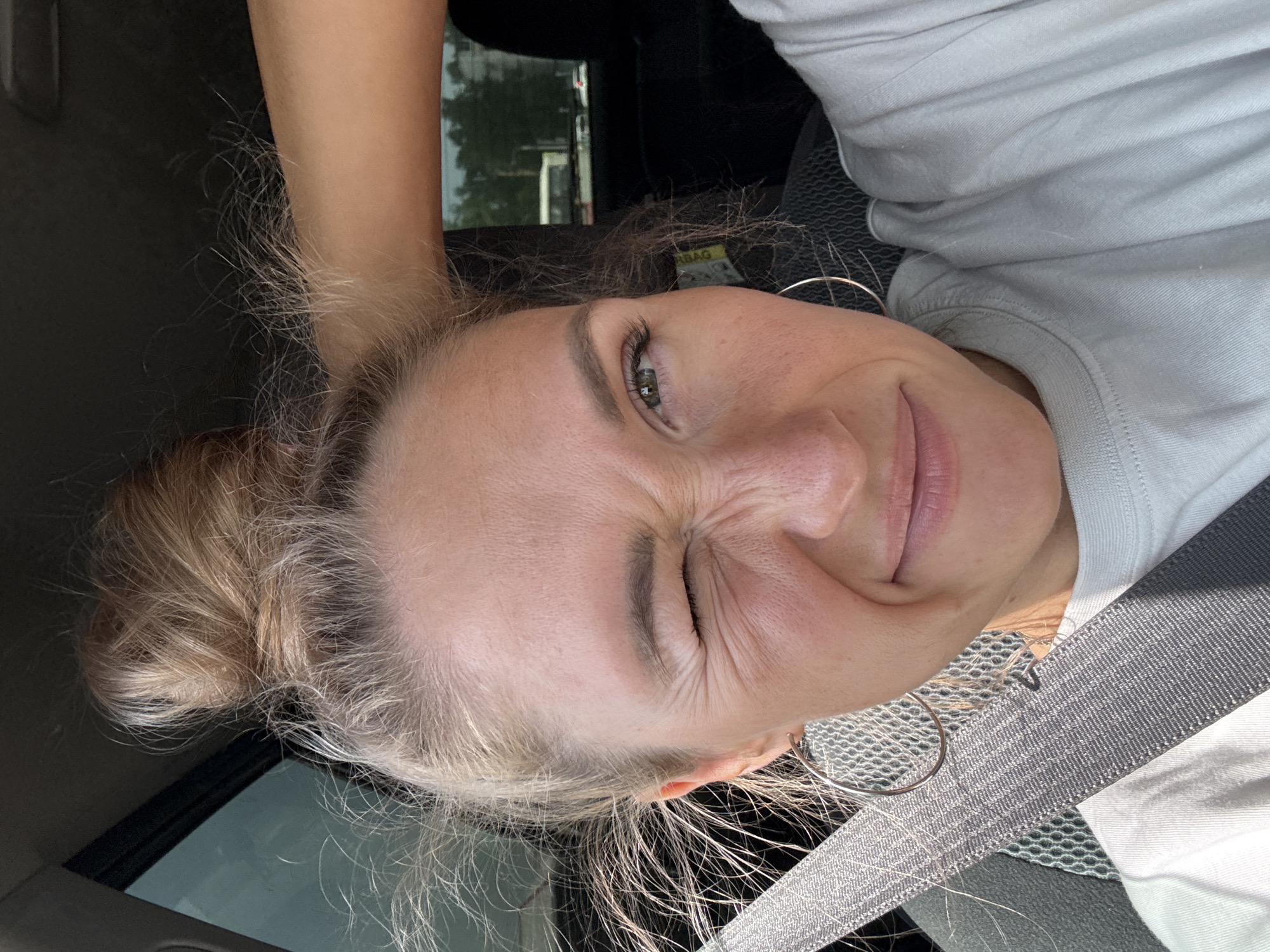 A selfie taken inside a car. The subject is a woman with blonde hair pulled back in a bun. She is wearing a white shirt and silver hoop earrings. A gray seatbelt is visible across her chest. She is winking one eye and smiling. The location cannot be determined.