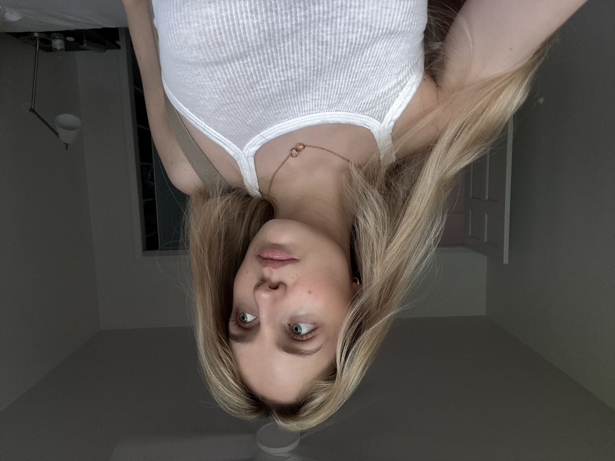 The image is a medium shot of a young woman with long blonde hair and light blue eyes, looking to her right. She is wearing a white ribbed tank top and a delicate gold necklace with two interlocking rings. The background appears to be an indoor setting, possibly a bedroom, with a gray wall on the left and a lighter colored wall on the right. A ceiling fan is visible at the top of the image. There is a doorway in the background on the left. On the right side of the background, a desk lamp with a white shade is on a dark surface, likely a nightstand, next to a bottle of water. The lighting in the room is dim, casting shadows and creating a muted atmosphere. The woman's skin has a light complexion, and she is wearing subtle makeup. The image is likely a selfie, given the angle.