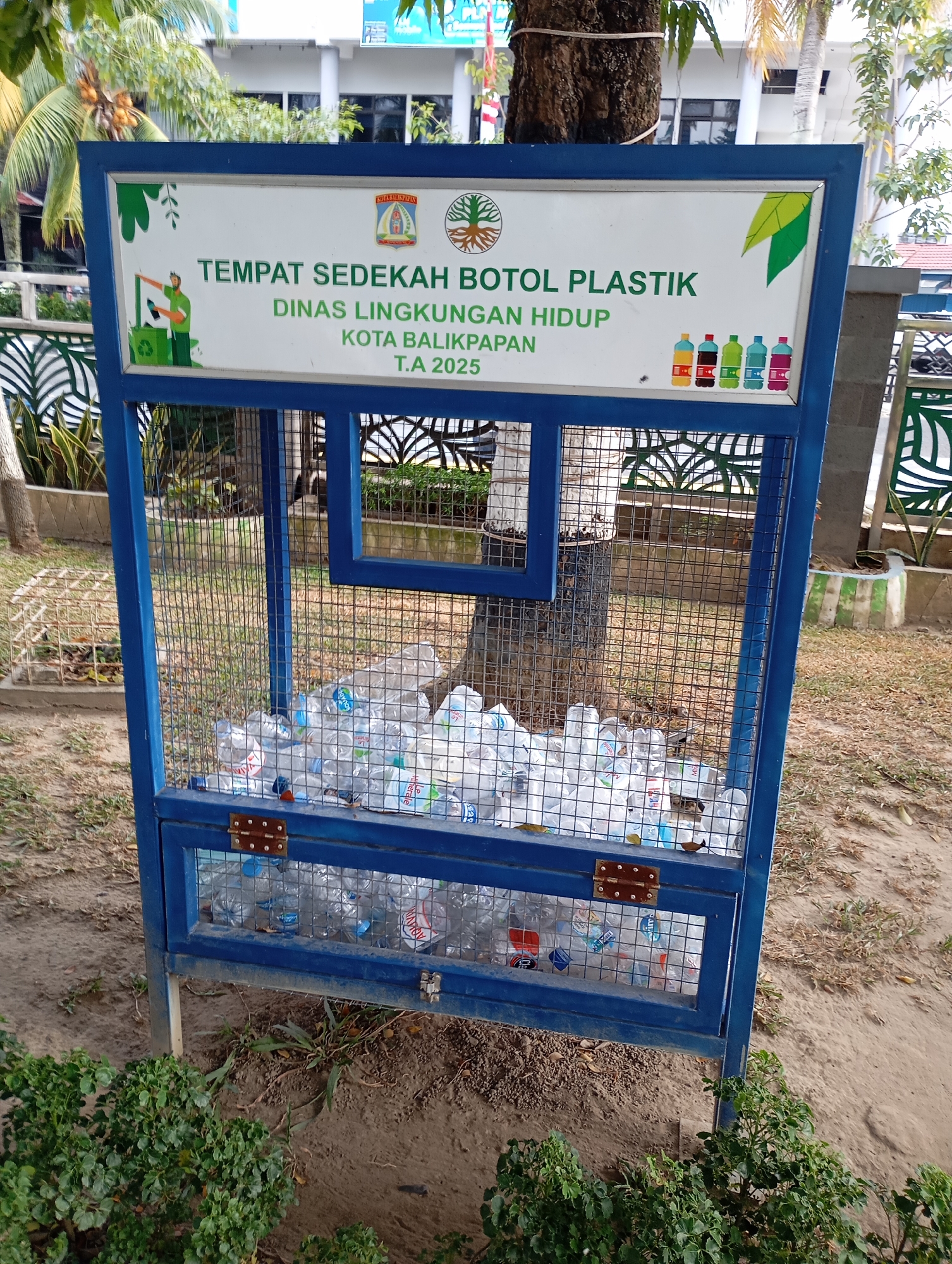 The image shows a blue metal recycling bin for plastic bottles. The bin has a sign at the top that reads "TEMPAT SEDEKAH BOTOL PLASTIK DINAS LINGKUNGAN HIDUP KOTA BALIKPAPAN T.A 2025". This translates to "Plastic Bottle Charity Place, Environmental Service, City of Balikpapan, Fiscal Year 2025". The bin is filled with plastic water bottles, some of which are clear and others are colored. The bin is located outdoors, in a grassy area with some trees and bushes. In the background, there is a building and a fence. The overall scene suggests a public space dedicated to environmental sustainability in Balikpapan, Indonesia.
