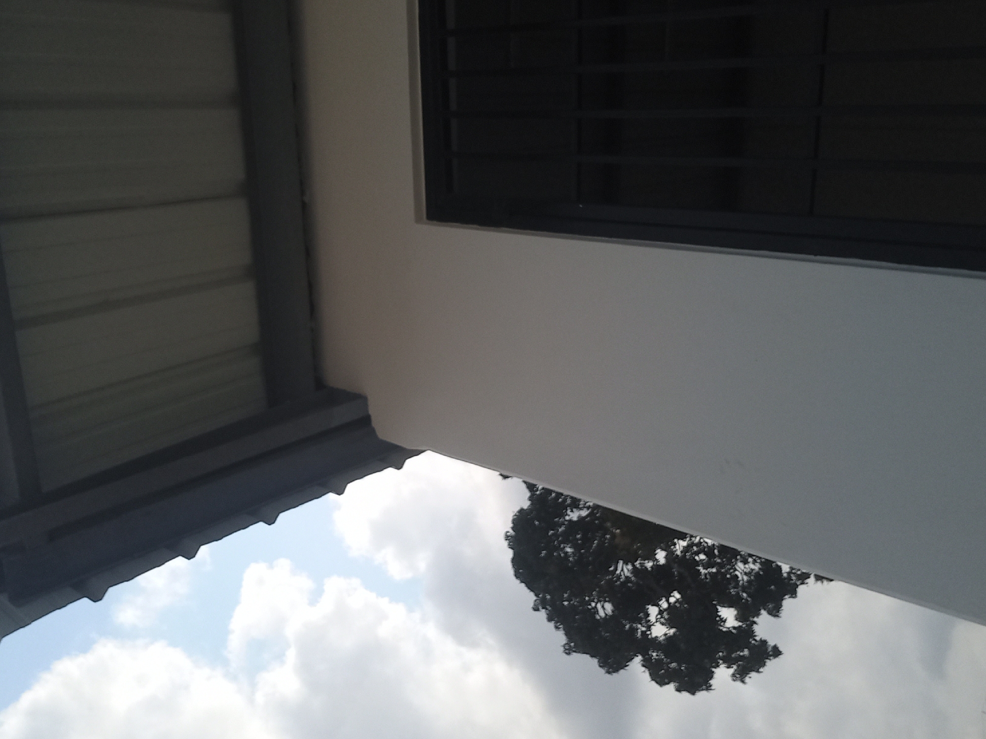 The image shows a close-up view of the exterior of a building in Bububu, Tanzania. A white wall is visible, with a dark-colored window grate on the right side. Above the wall, a gray metal roof with ribbed panels is present, extending diagonally across the upper portion of the frame. In the background, a large tree with dark foliage is seen against a cloudy sky. The lighting suggests it is daytime. The scene is devoid of people or any discernible activity.