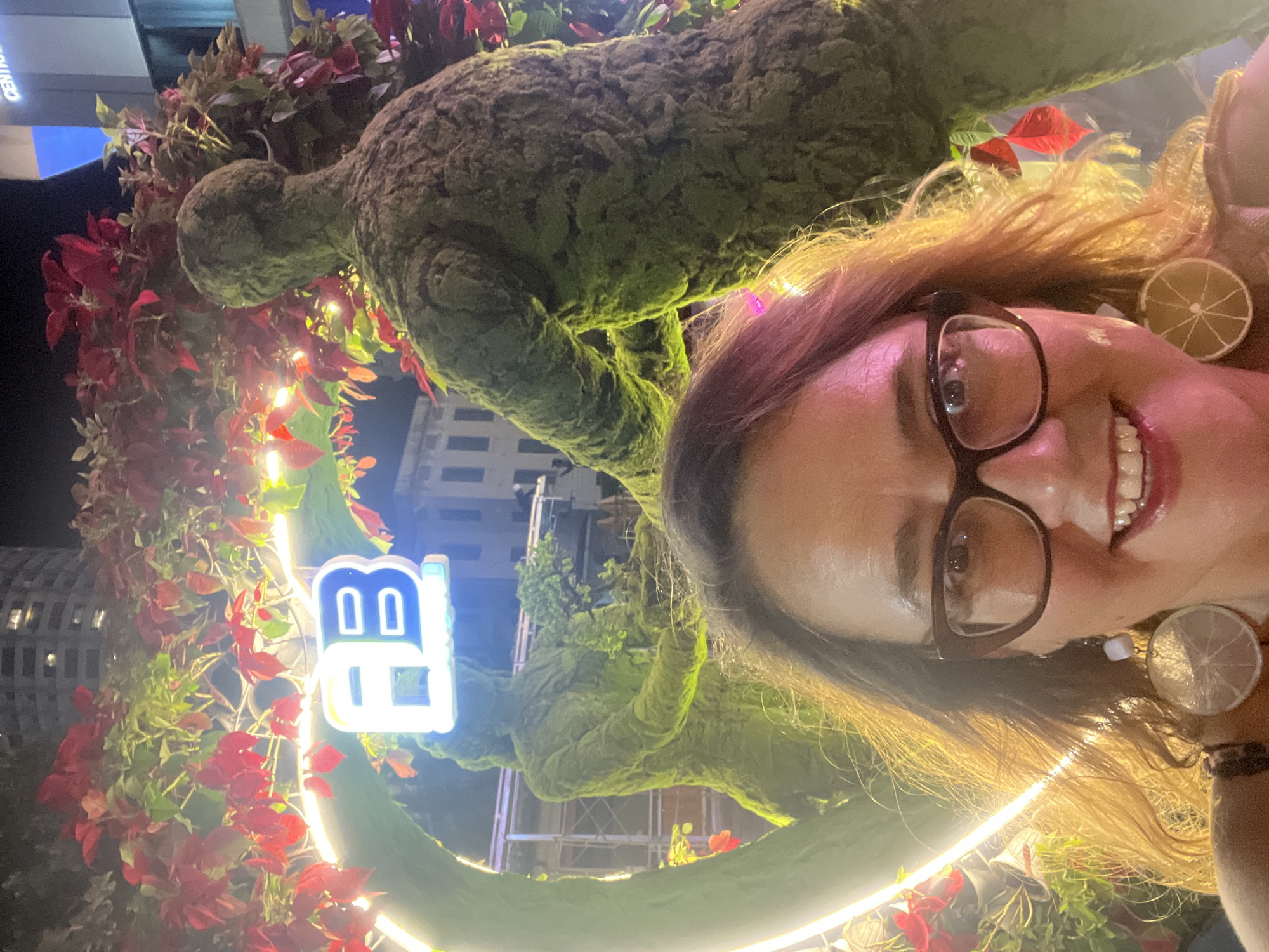 In Nha Trang Ward, Vietnam, a woman with glasses and curly brown hair poses for a selfie in front of an outdoor art installation. The installation features two human-shaped sculptures covered in green moss, with red and green foliage overhead. A glowing neon "FB" sign is visible between the sculptures, which appear to be intertwined with a large, illuminated ring. The woman is wearing large, circular earrings with a citrus slice design. The background shows a dimly lit urban environment with building structures.