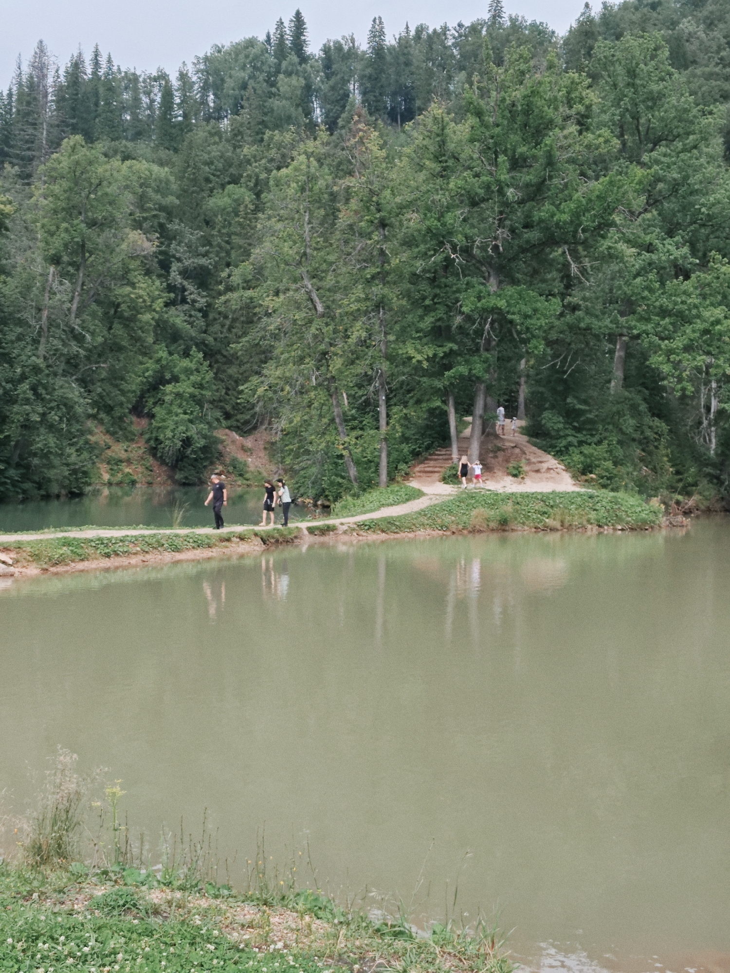 A wide shot captures a tranquil lake in Krasnyy Klyuch, Russia, bordered by a dense, green forest. A narrow, sandy path winds along the edge of the water, with several people walking on it. In the foreground, the lake's surface is calm, reflecting the surrounding greenery and the overcast sky. On the path, three individuals are visible in the mid-ground: one person wearing a black t-shirt and dark pants, another in a black dress, and a third in a light blue shirt and dark pants. Further along the path, closer to the forested area, a smaller group of people can be seen, including a person in a light-colored shirt and dark shorts, and two children. The forest is lush and dense, with a mix of deciduous and coniferous trees, extending up a gentle slope from the lake. The path itself appears to be a popular walking or hiking trail, with some steps leading up a small incline in the distance. The overall atmosphere is serene and natural.