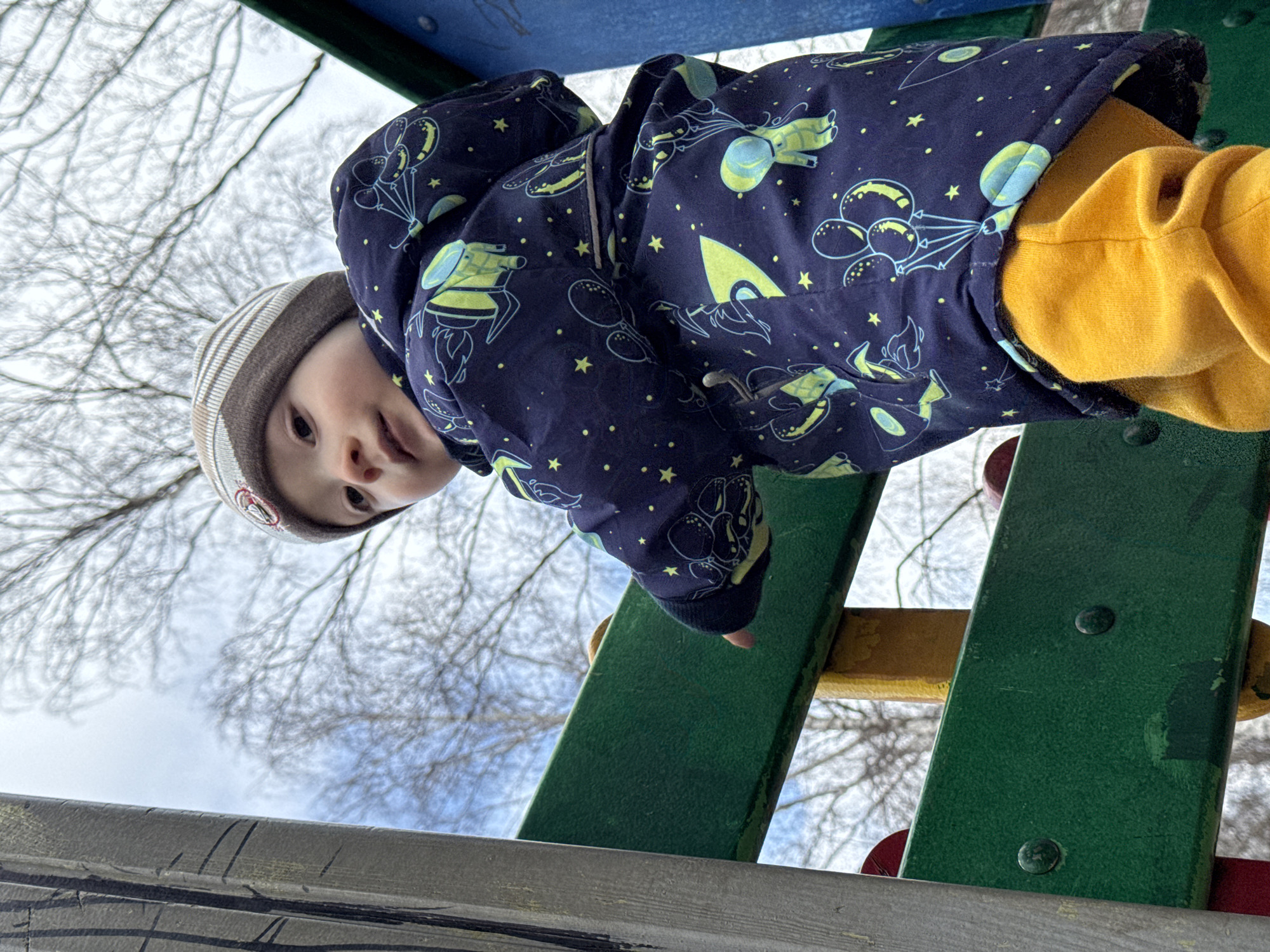 The image presents a low-angle perspective looking up at a young child from below a playground structure. The child, appearing to be a toddler, is positioned above the camera, peering down. They are dressed in a dark blue jacket adorned with light blue and yellow patterns of stars, balloons, and rockets, paired with bright yellow pants. A striped beanie in muted tones of beige and brown covers their head. The child's face is visible, with dark eyes looking directly forward, exhibiting a curious or mild expression.
The child's left hand rests on a green-painted beam of the playground equipment. Other components of the structure, including yellow and potentially red elements, are partially visible around the child, suggesting a colorful outdoor play area. In the background, stark, bare tree branches are silhouetted against a bright, overcast sky. The leafless trees indicate a season of late autumn, winter, or early spring. The lighting suggests it is daytime. No visible text can be discerned in the image. The scene is set in Izhevsk, Russia.