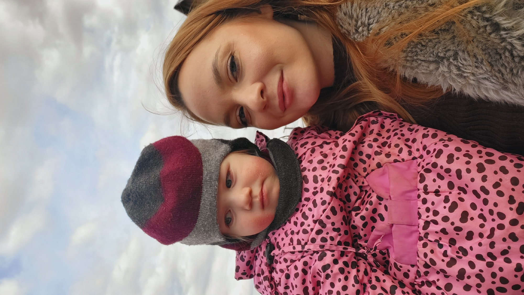 The image displays a close-up, upward-angled portrait of a young woman and a baby or toddler outdoors in Зипуново, Russia. The background features a pale, cloudy sky.
The woman is positioned in the upper part of the frame. She has long, reddish-brown hair and a gentle, subtle smile, looking down towards the camera. She appears to be wearing a grey, possibly faux fur, collar or coat.
Below her, the baby or toddler looks towards the camera with a neutral expression. The child is dressed in a vibrant pink winter jacket adorned with black leopard-like spots. A warm, knitted hat in shades of grey, maroon, and white covers the child's head. The child's cheeks have a rosy tint, indicative of cold weather.
The overall scene suggests a cool or cold daytime environment, given the subjects' winter attire and the cloudy sky. The photograph seems to be a selfie taken from a low angle, capturing a moment between two individuals, likely a parent and child. No text is visible in the image.