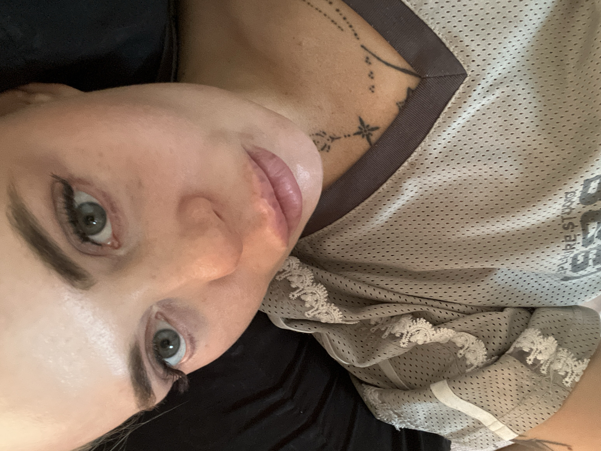 An overhead close-up, eye-level shot shows a Caucasian woman's face, taking up most of the frame, with her head tilted slightly to her left. She has fair skin, prominent cheekbones, light brown eyebrows, and light grey-green eyes with dark mascara and false eyelashes. Her lips are slightly parted and appear plump, with a subtle sheen of lip gloss. She is wearing a grey, mesh athletic shirt with white lace trim on the left shoulder and sleeve. A dark brown V-neck collar is visible. A portion of her left arm, bent at the elbow, is visible in the lower left of the frame. She has a small tattoo on her collarbone, featuring a star with lines extending from its points and a floral design above it. The background is dark and out of focus, with hints of black and brown fabric, possibly a bed or couch. The lighting is warm and diffused, casting soft shadows on her face and highlighting the texture of her skin.