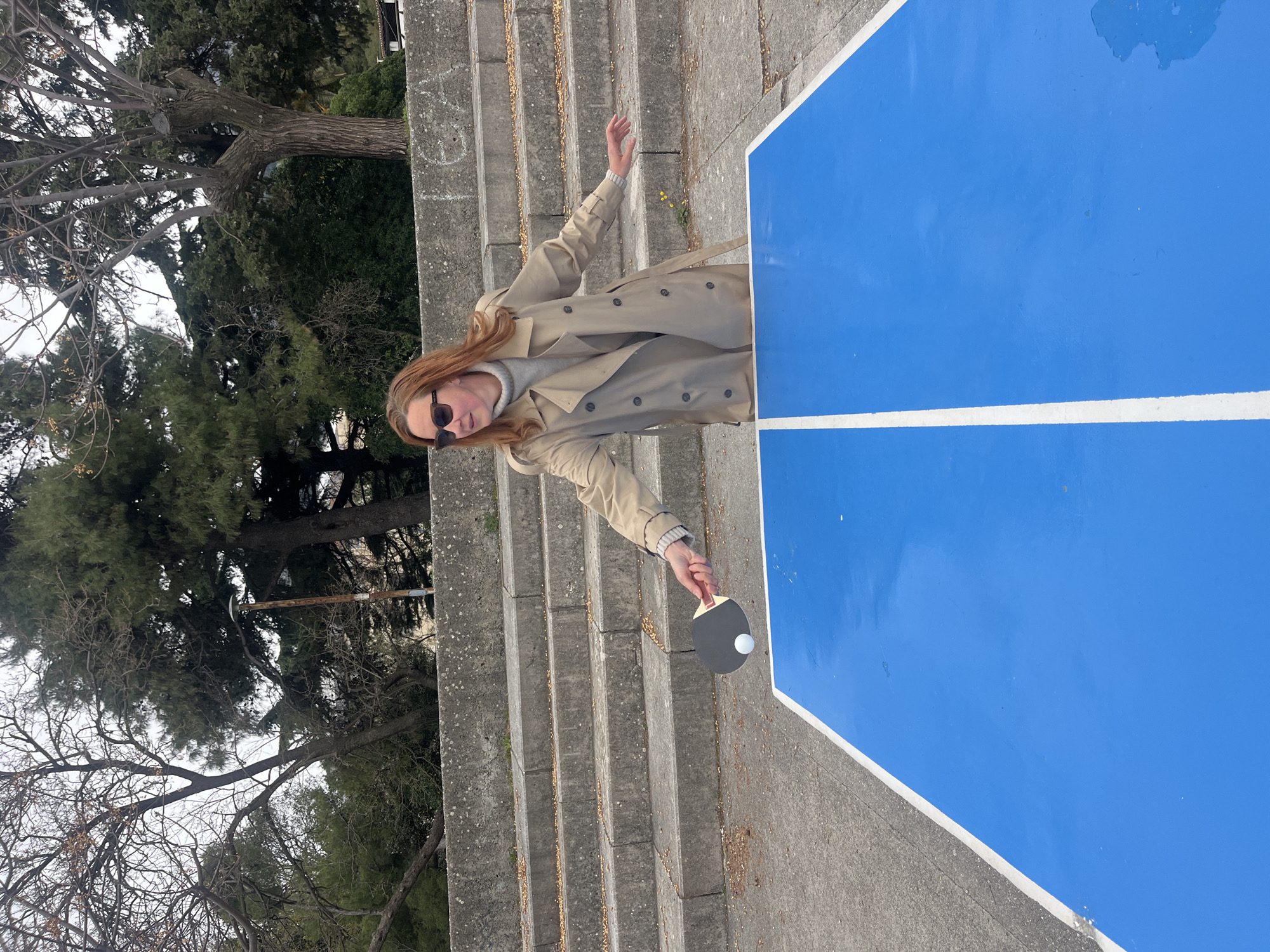 A medium outdoor shot shows a woman playing ping pong in Budva, Montenegro. She stands on concrete steps leading up to the left of the blue ping pong table. She is light-skinned with long red hair and wears a light-colored trench coat over a gray turtleneck, and dark sunglasses. The ping pong paddle in her right hand is black and gray, with a small white ball between it and the table. Her left hand is raised out to her side. The steps behind her are concrete, and some leaves or small brown material is scattered on them. A tree is visible behind the steps. The blue ping pong table with a white stripe is off to the right. The sky is overcast.